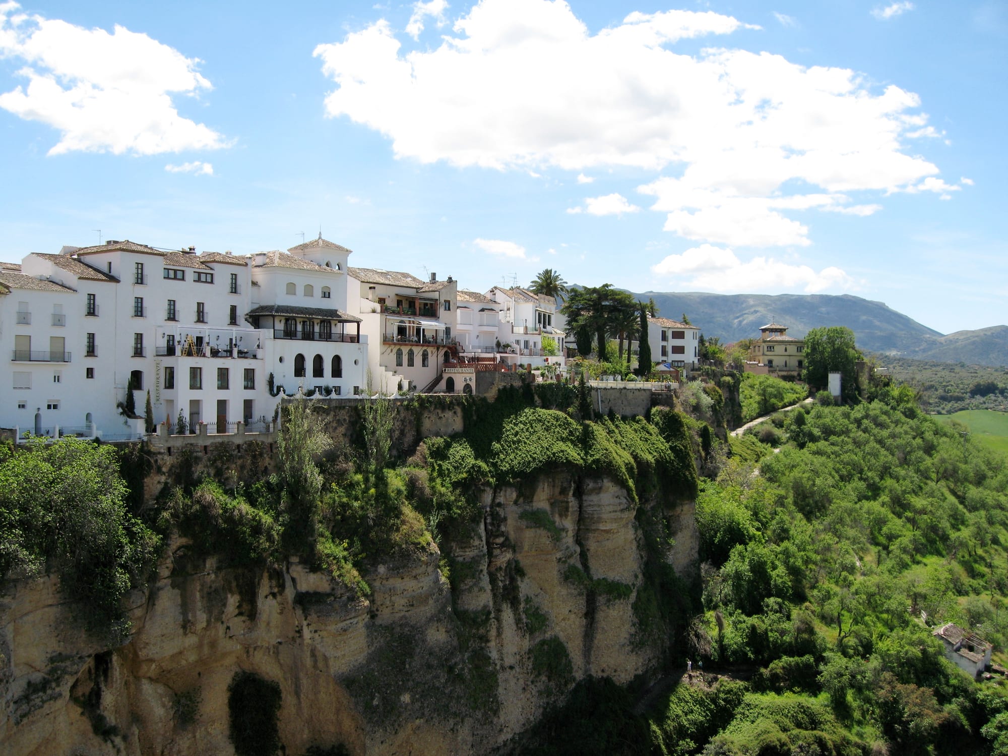 White houses with balconies lining the edge of a sheer, vegetation-covered cliff, with green hills and mountains beyond