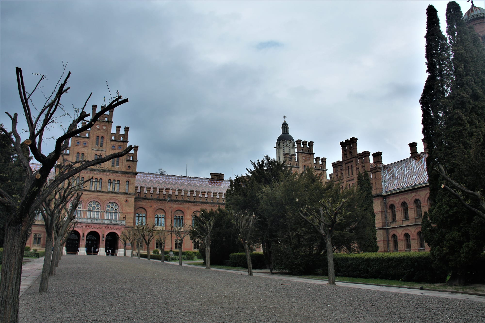 Red-brick buildings with crenellated towers and patterned tile roofs around a gravel courtyard with trees, under grey clouds