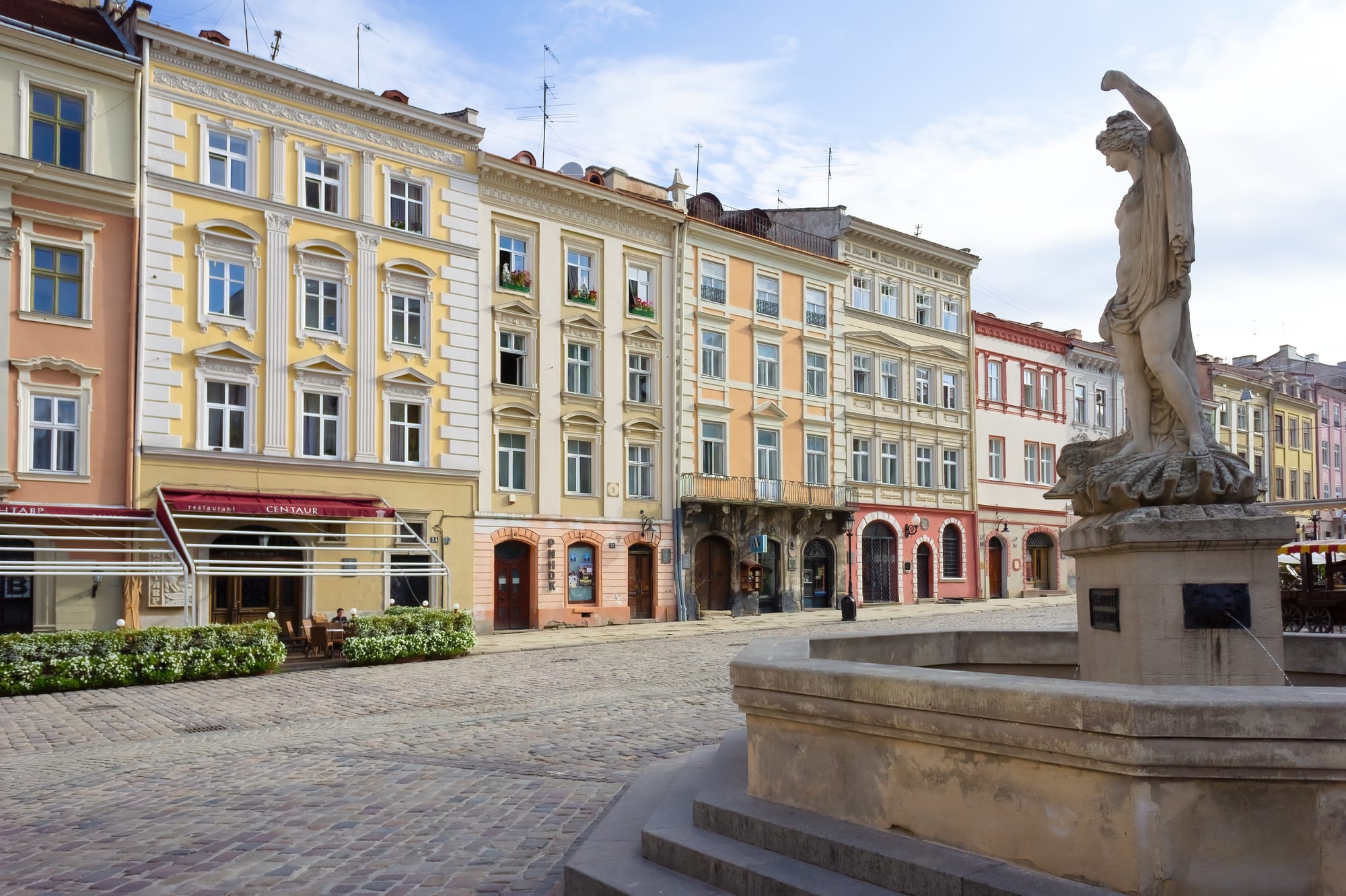 Pastel merchant houses on a cobbled square in Lviv, with a stone statue of a sea goddess on a fountain in the foreground