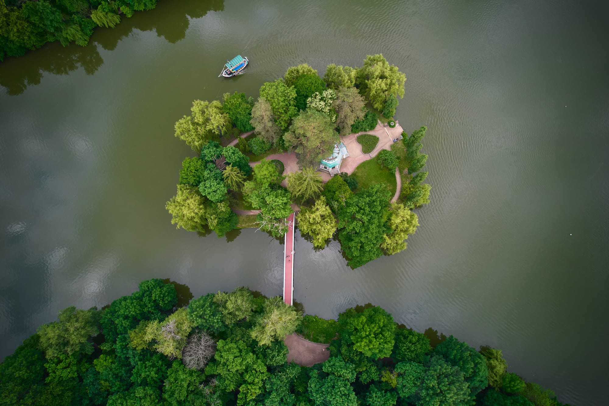 Aerial view of a small wooded island with a pavilion and paths, joined to the shore by a footbridge across a green lake