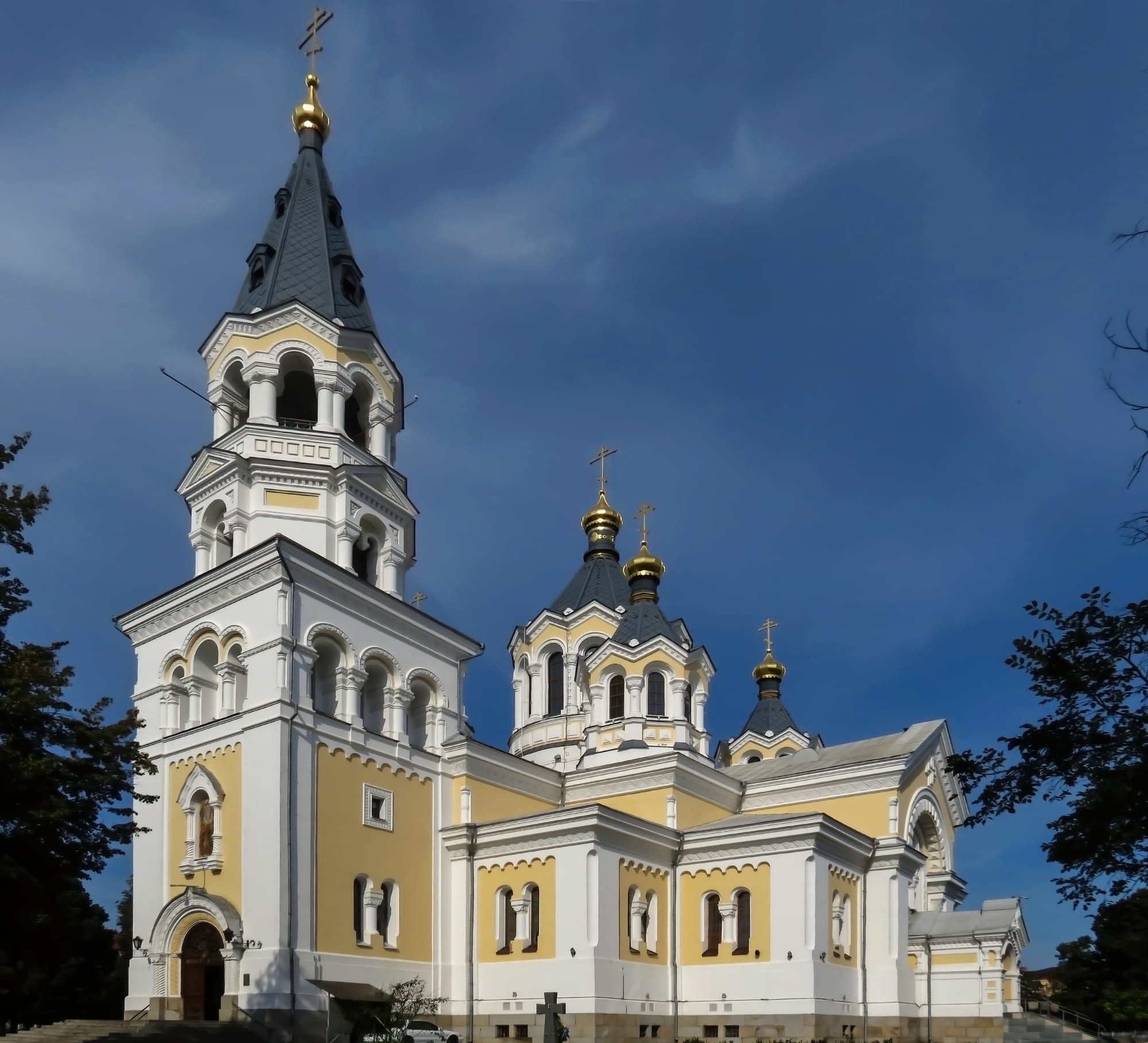 A cream and white Orthodox cathedral with a tall bell tower and dark domes topped with gold, under a deep blue sky