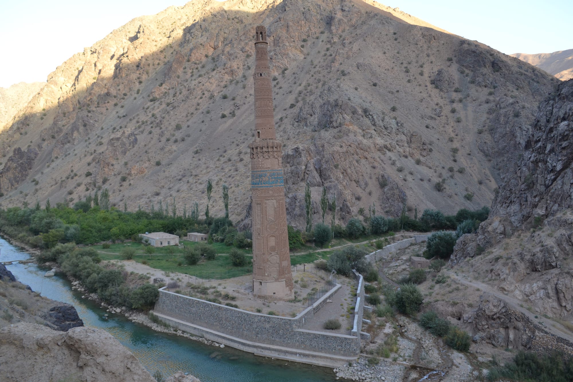 A tall, slender brick minaret with a blue tiled band standing beside a green river below bare brown mountains