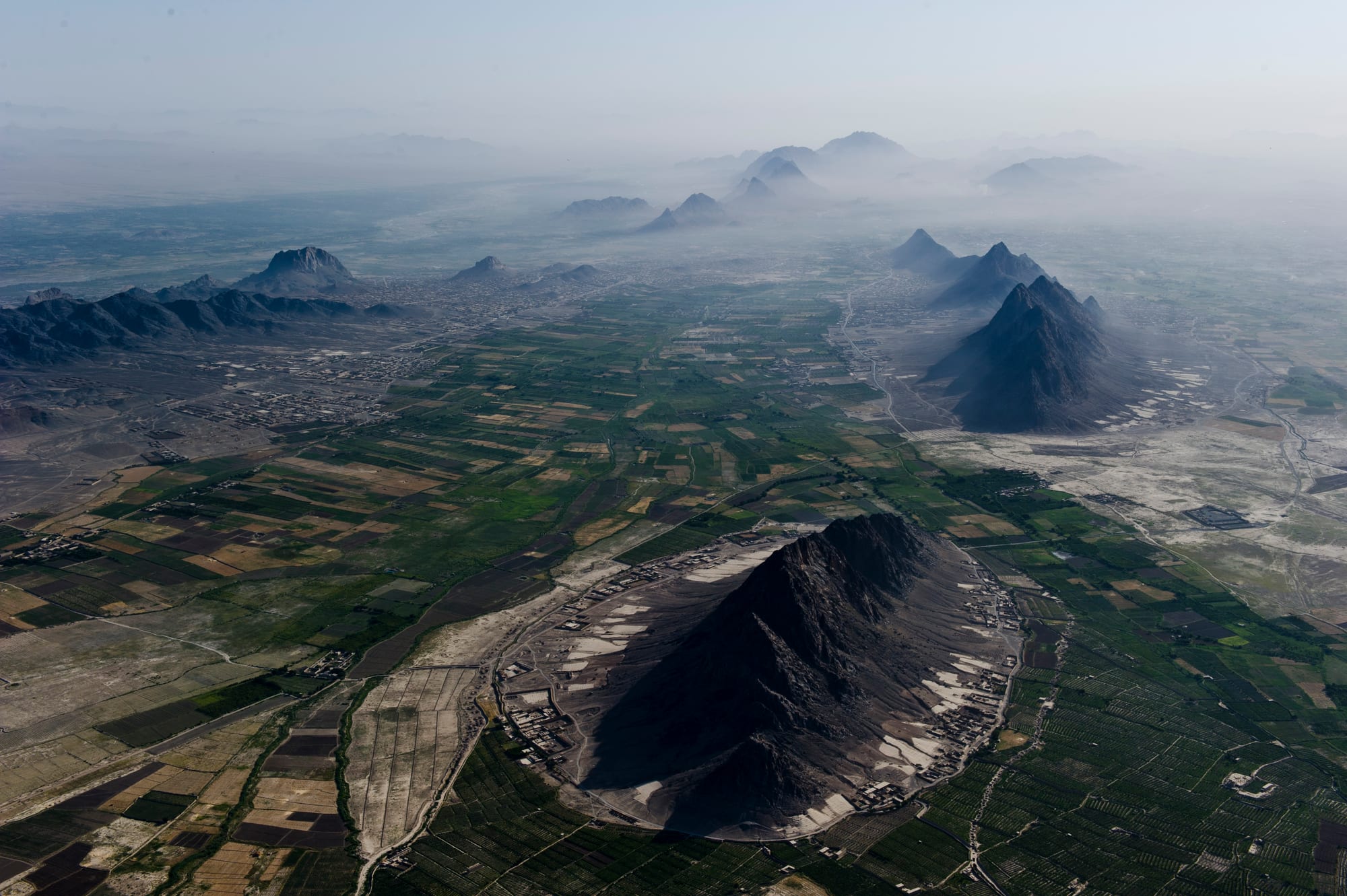 Aerial view of green irrigated fields and villages around dark, steep rocky hills fading into a hazy plain