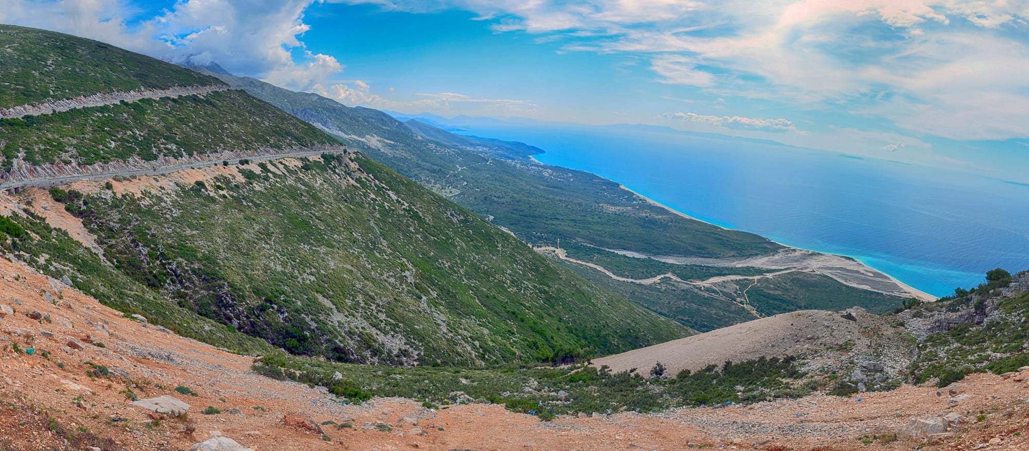 Green mountainside with a road cut across it, dropping to a long pale beach and the bright blue Ionian Sea