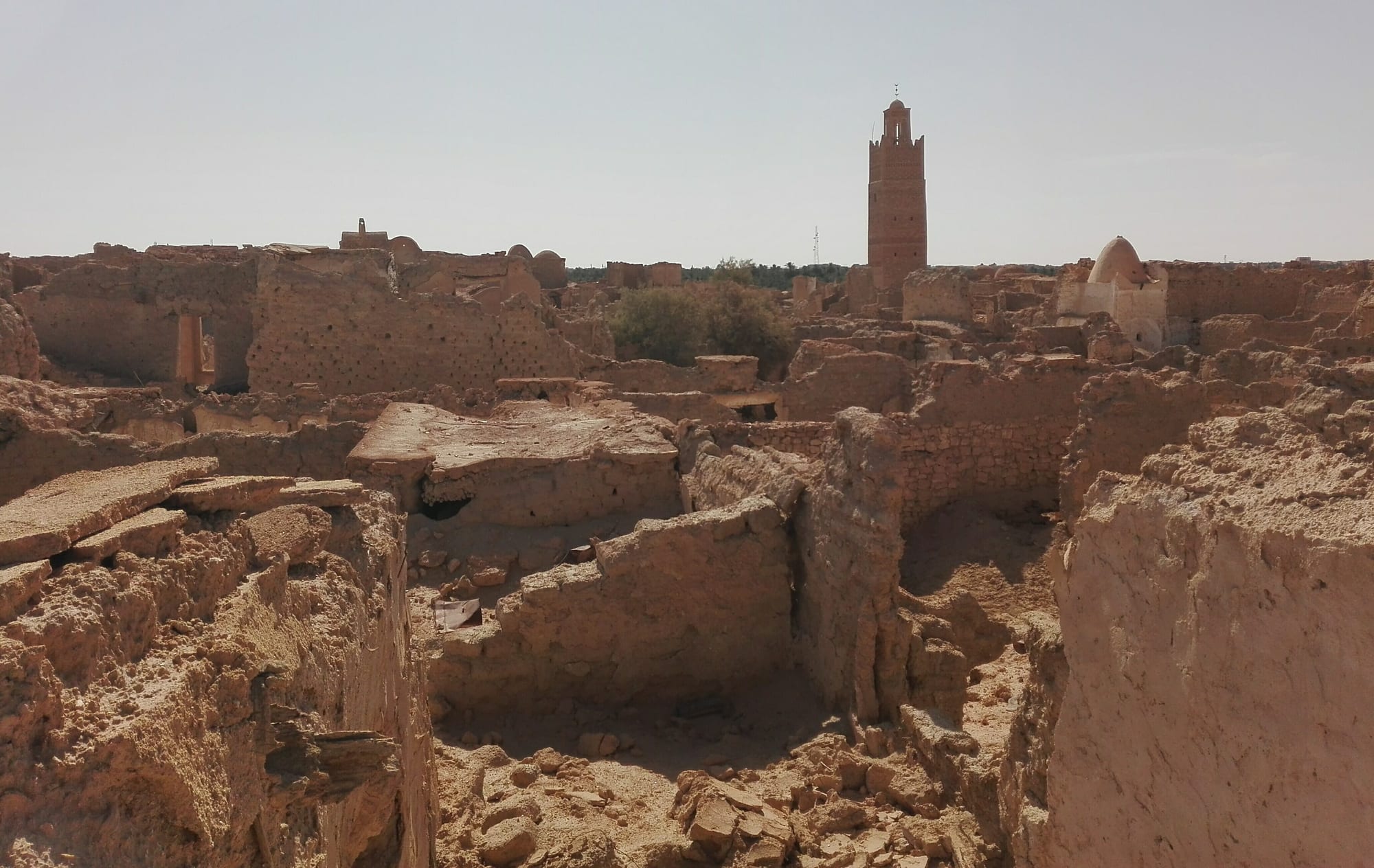 Crumbling mud-brick walls and roofless houses of an old desert village, with a tall brick minaret and a white dome rising behind under a hazy sky