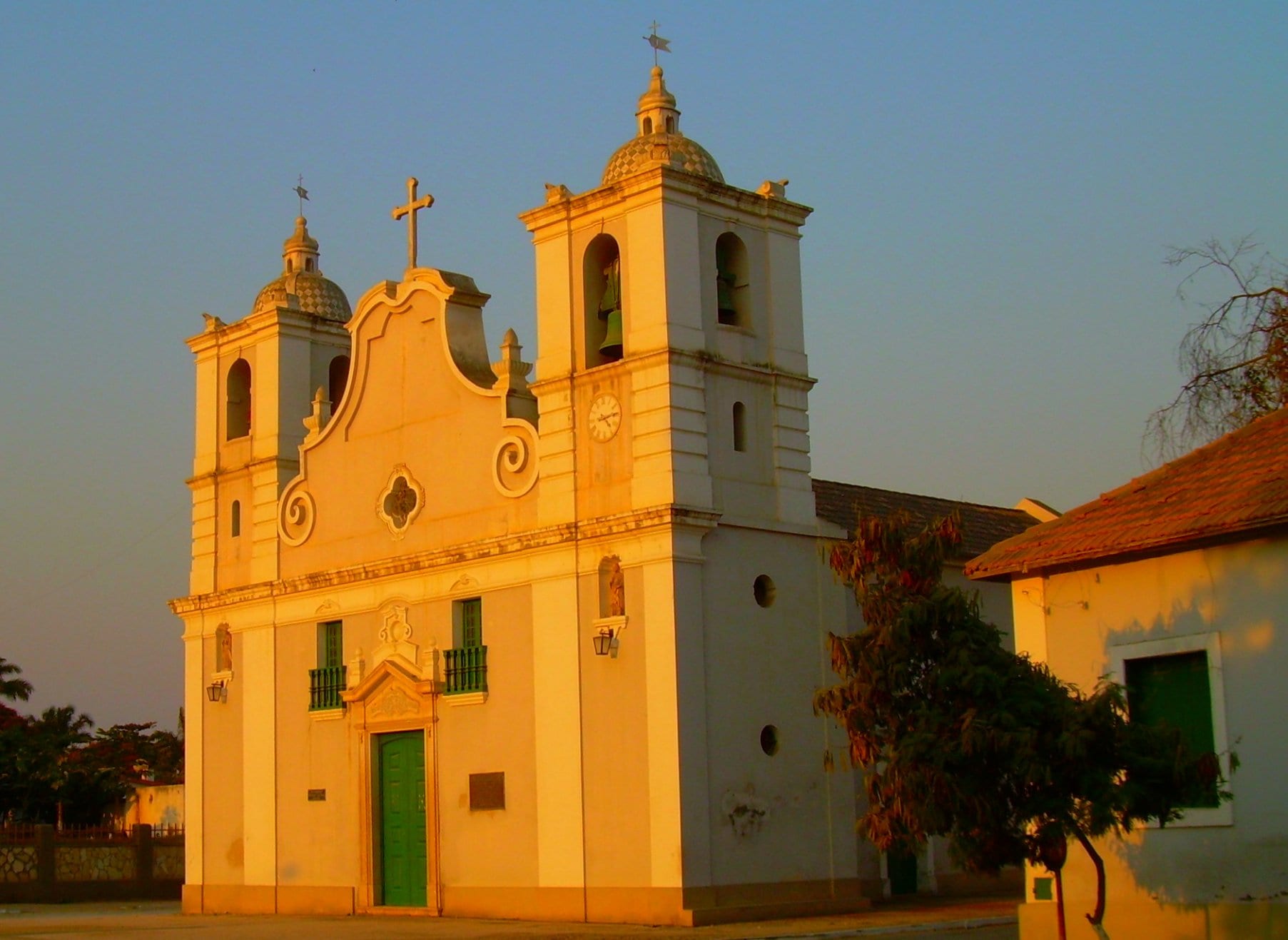 A white colonial church with two bell towers and a curved gable, glowing in warm evening light under a clear sky