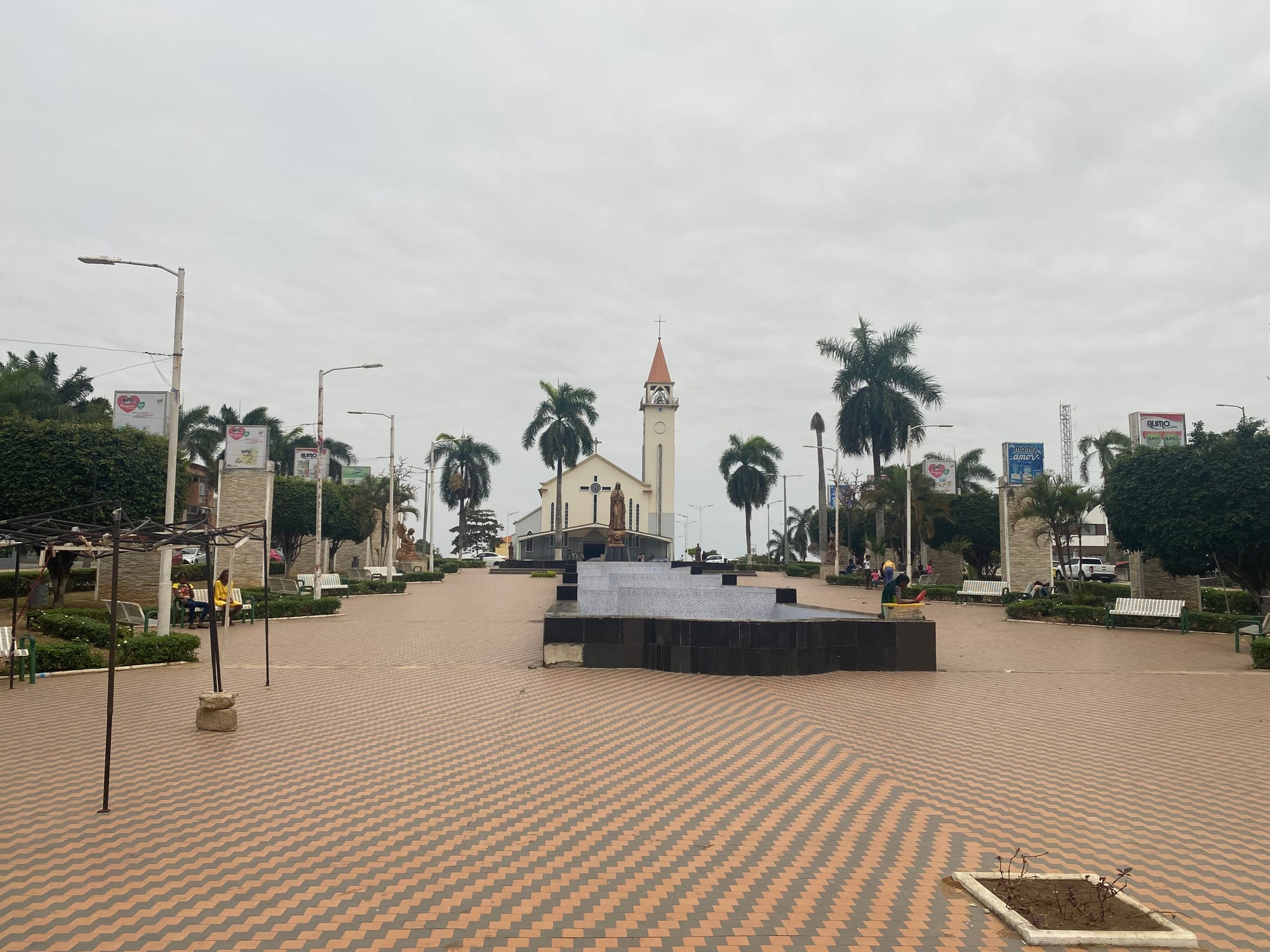 A paved square with a fountain, benches and palm trees leading to a white church with a tall bell tower