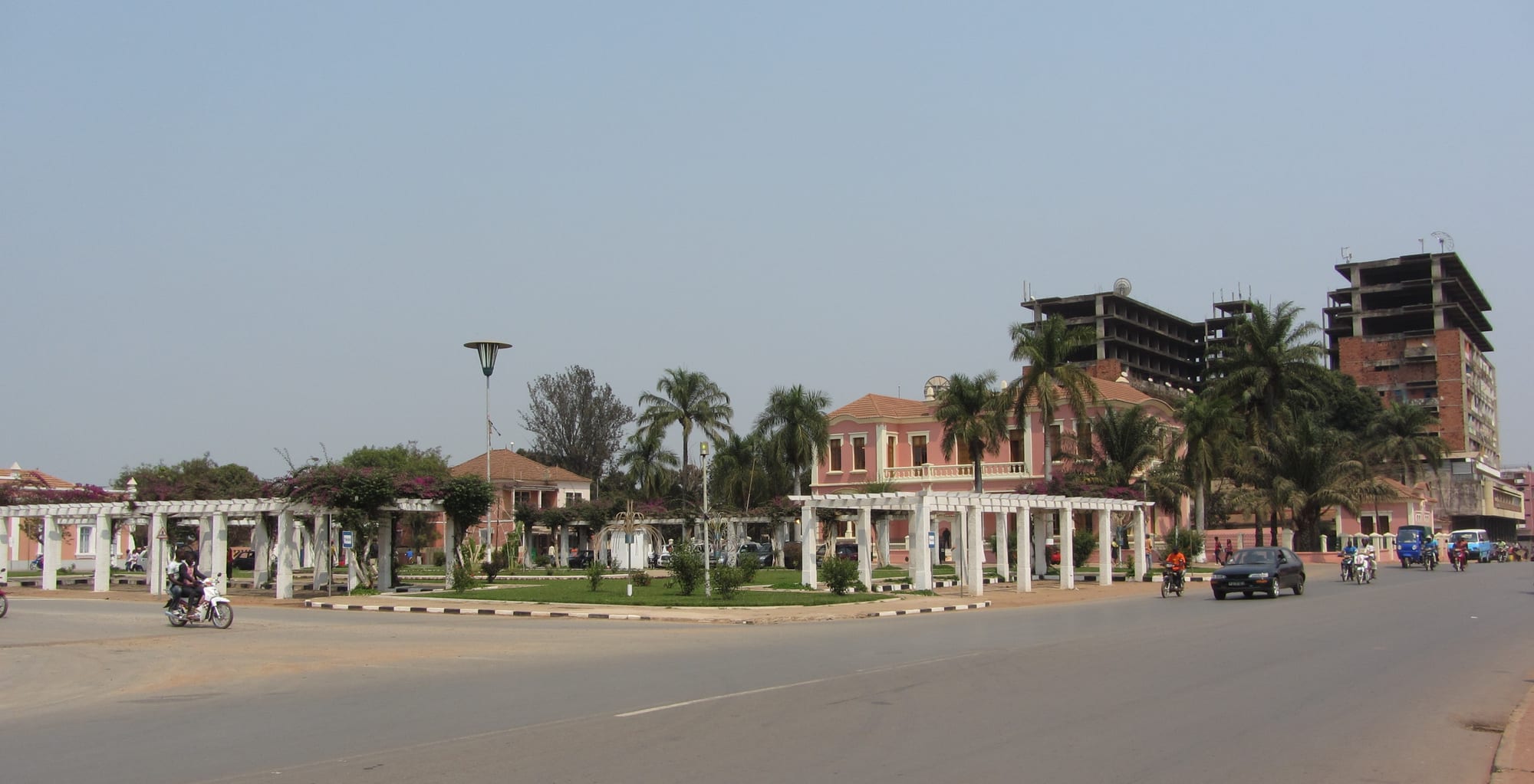 A wide street curving past a small garden with white pergolas, palm trees and pink colonial buildings, with taller concrete blocks behind