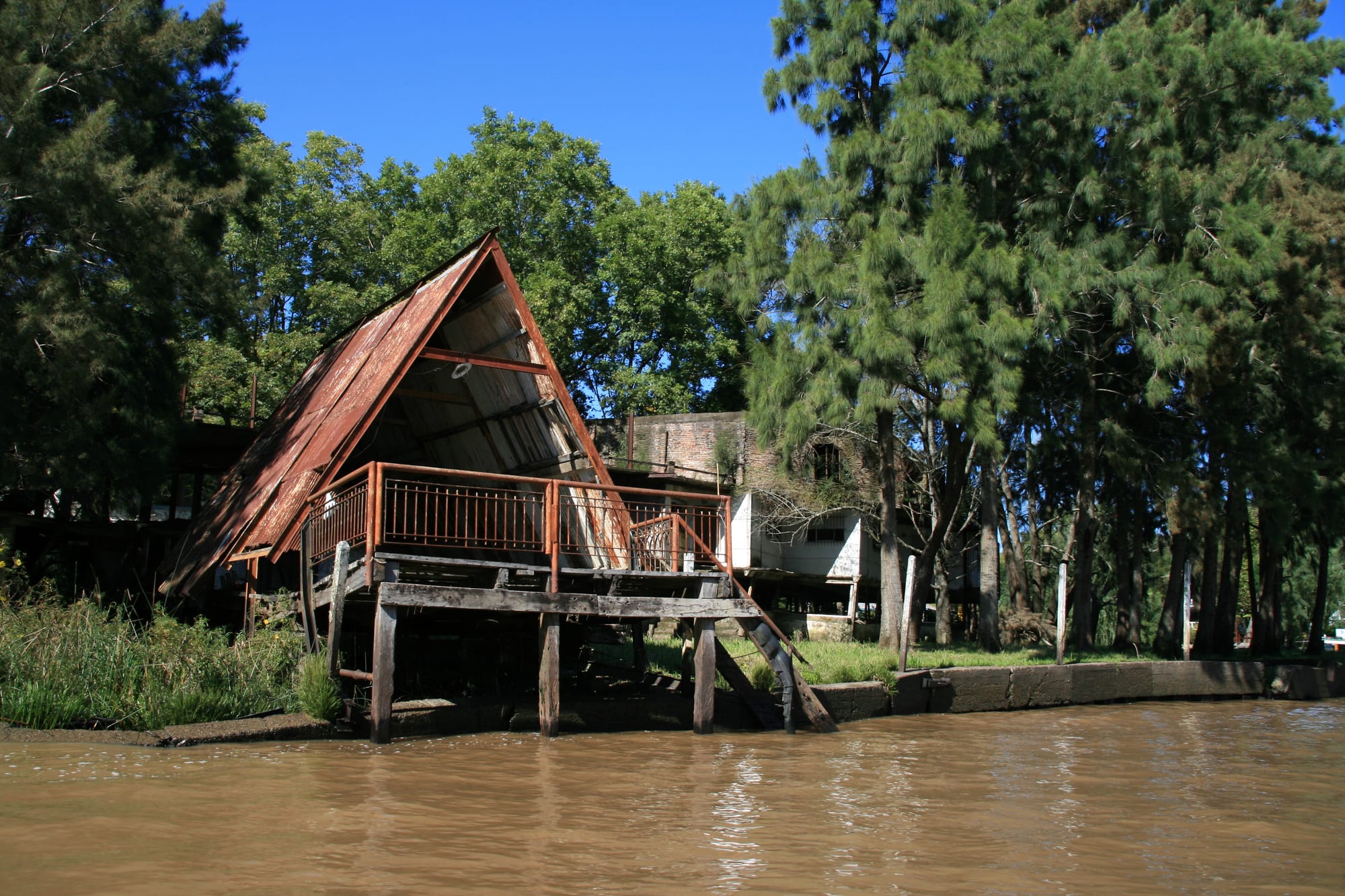 An A-framed wooden boat shed on stilts over a brown river channel, backed by tall trees under a clear blue sky