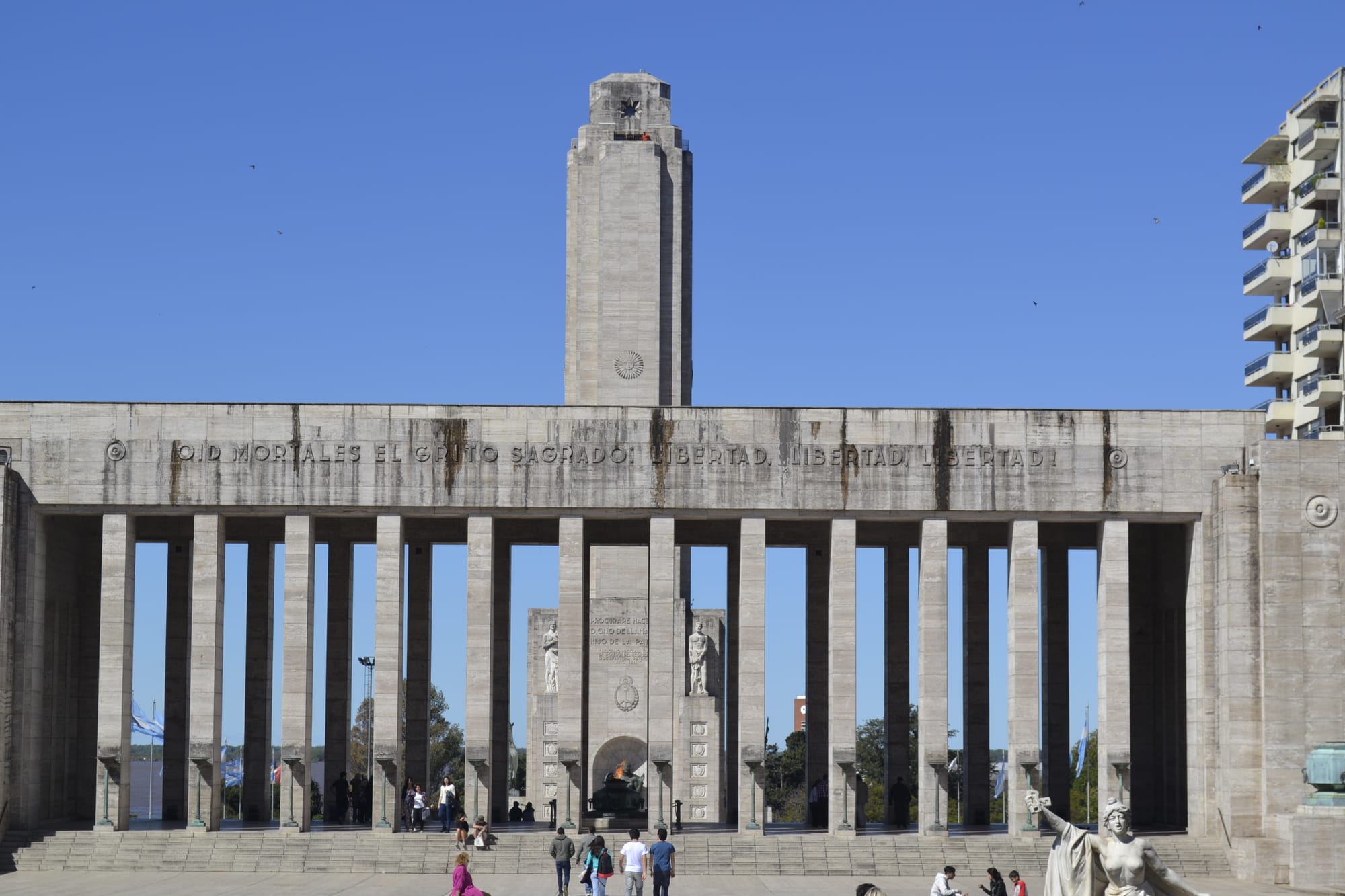 A long stone colonnade inscribed with the national anthem and a tall tower behind it under a clear blue sky