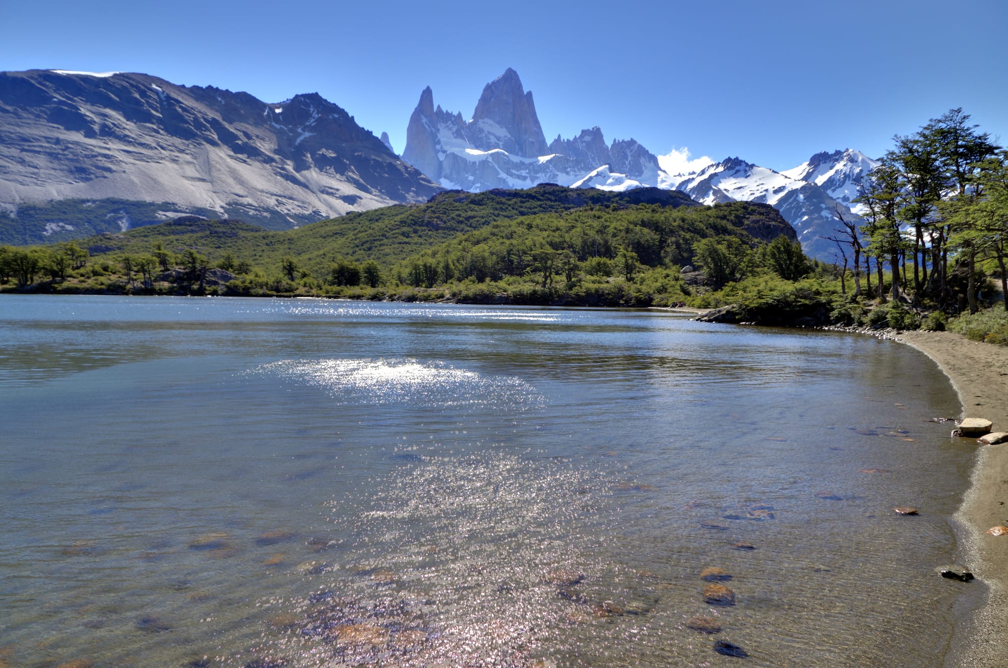 Jagged snow-streaked granite peaks rising above wooded hills and a calm, clear lake with a pebble shore