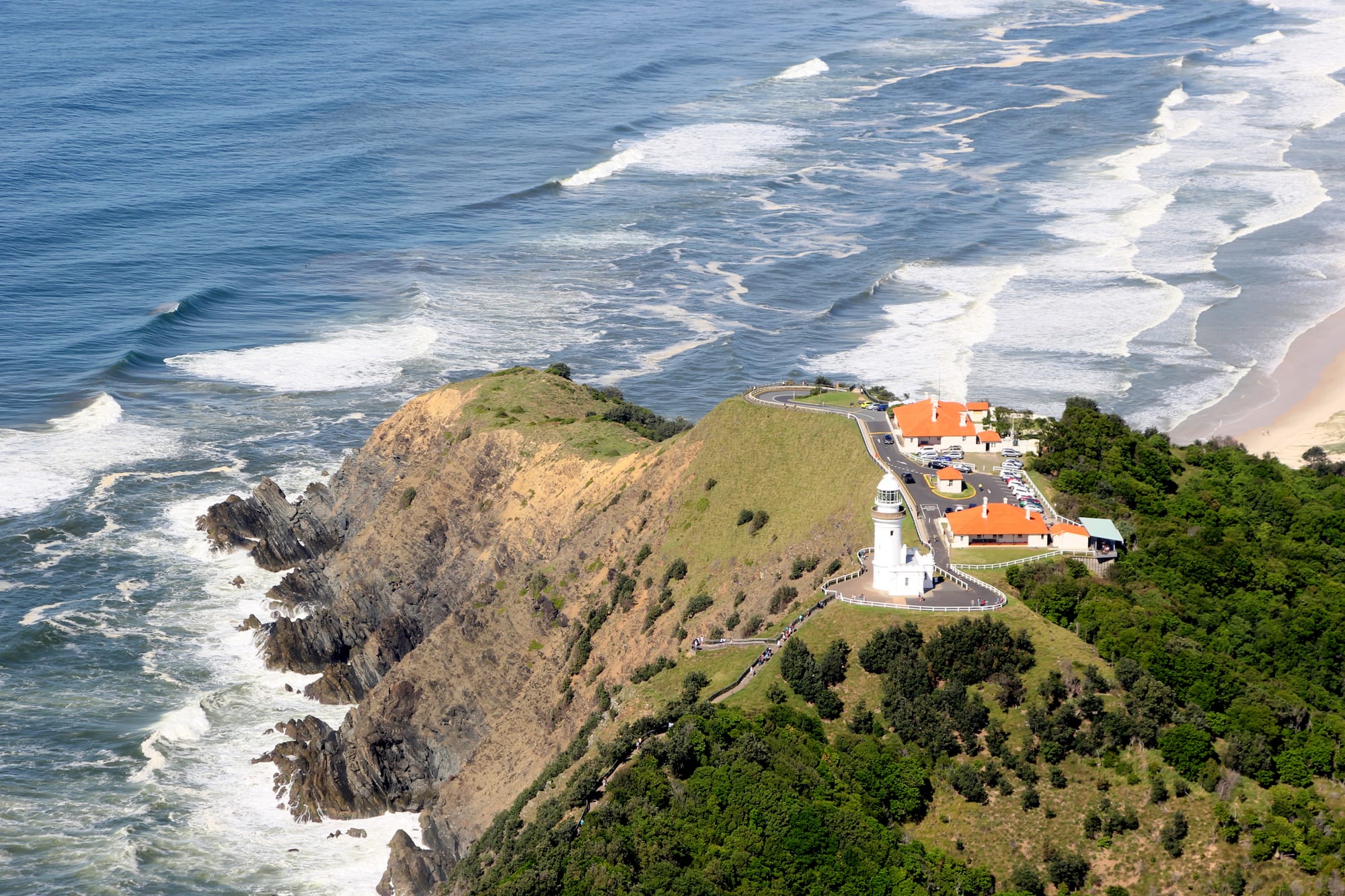 Aerial view of a white lighthouse on a green headland above rocky cliffs, with surf rolling onto a sandy beach