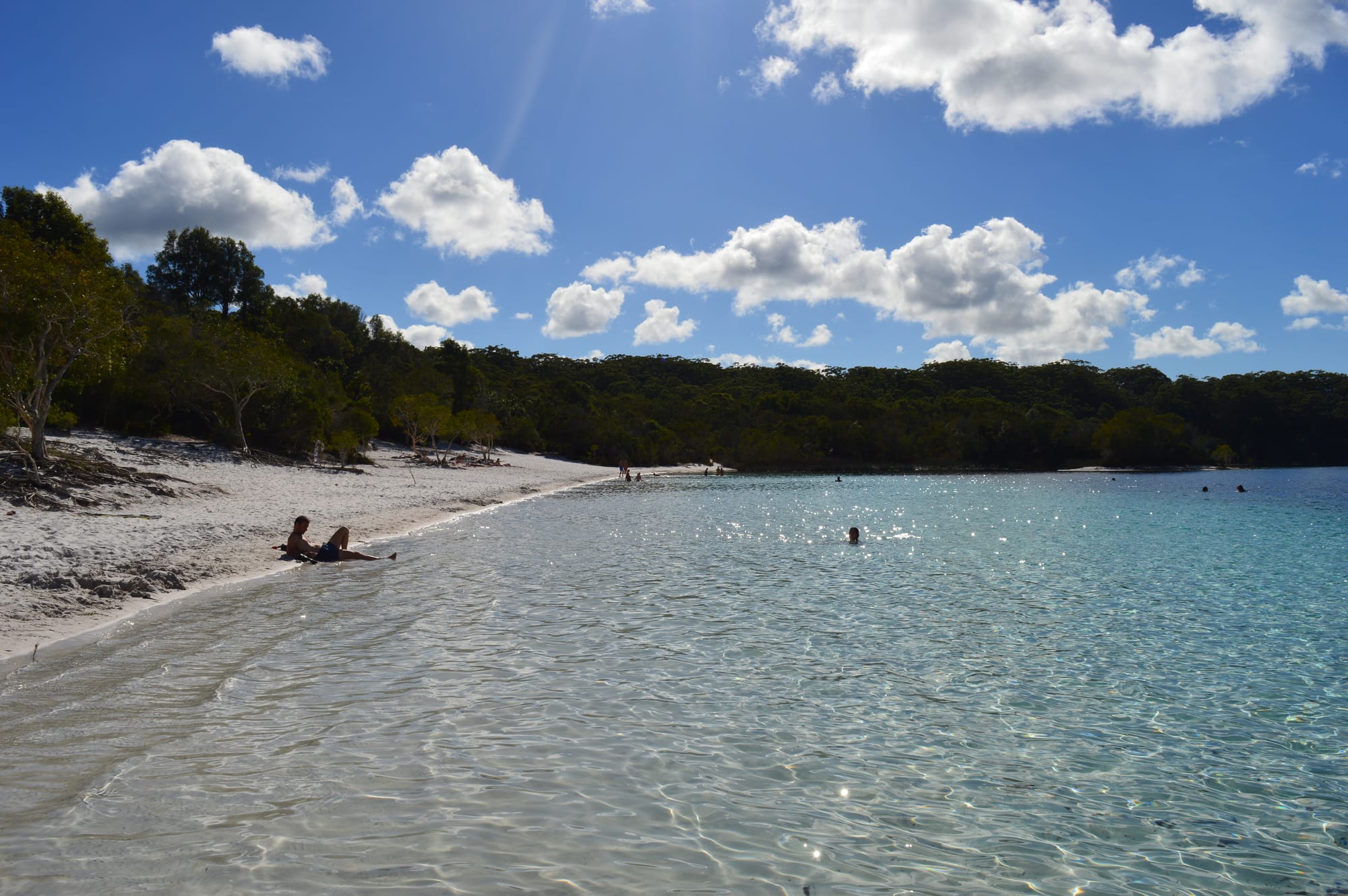 Clear turquoise freshwater lapping a white sand shore, with eucalypt forest around the lake under a cloudy blue sky