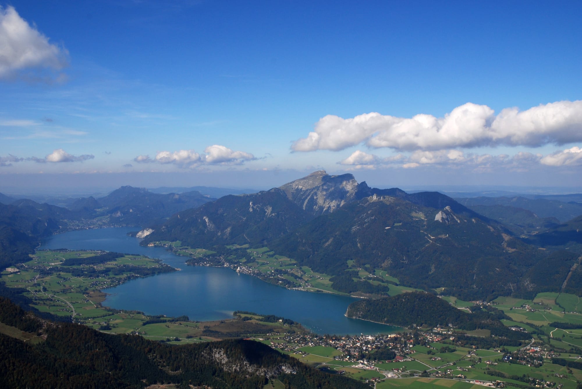 Distant view of a long blue lake among green meadows and villages, with a rocky mountain rising above its far shore