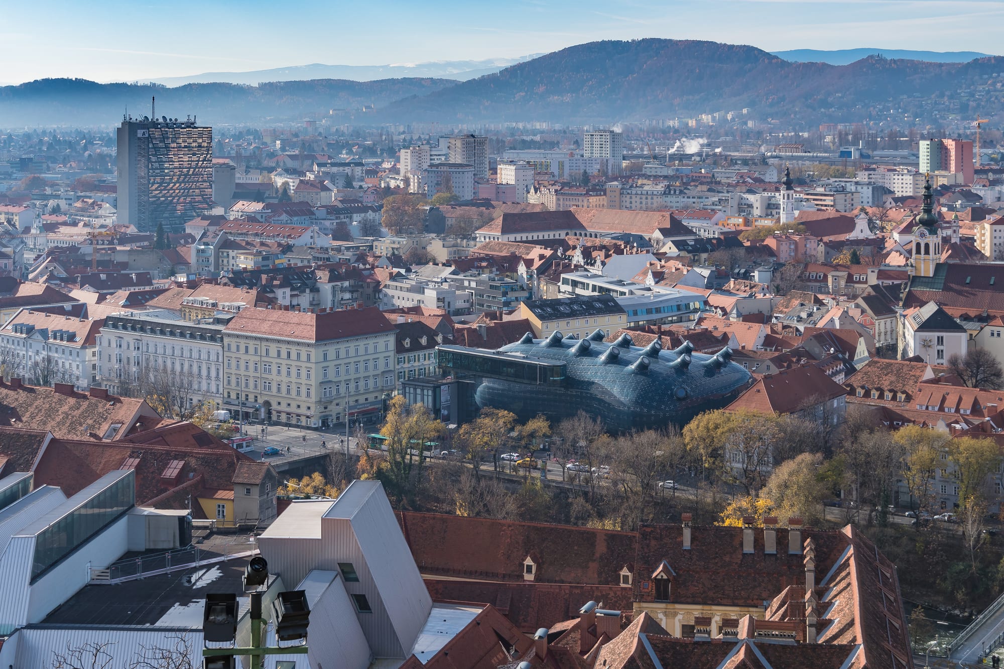 Red-tiled rooftops of Graz seen from above, with the blue curved Kunsthaus in the centre and wooded hills beyond