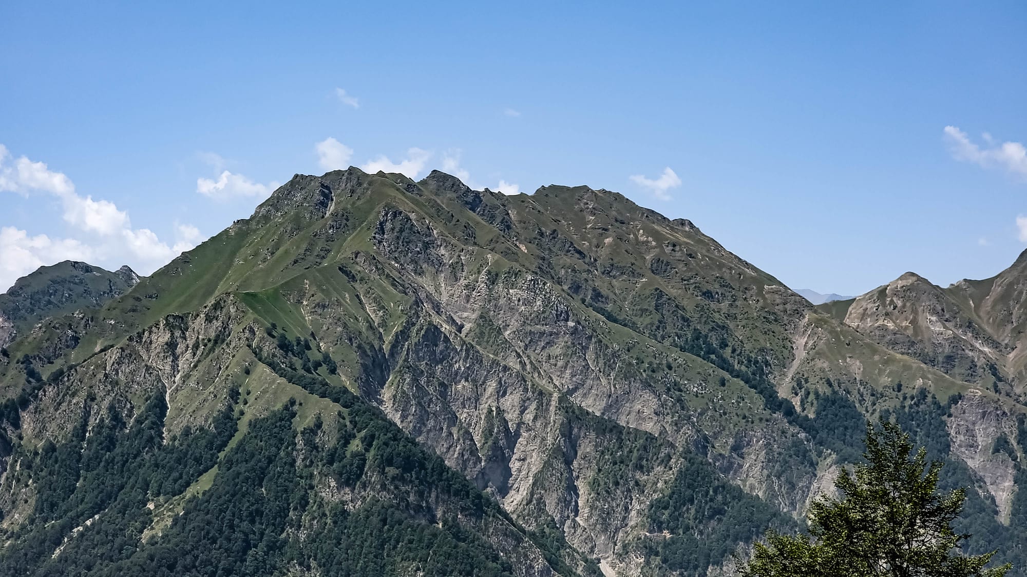 A rugged green mountain with rocky cliffs and forested lower slopes under a blue sky with a few small clouds