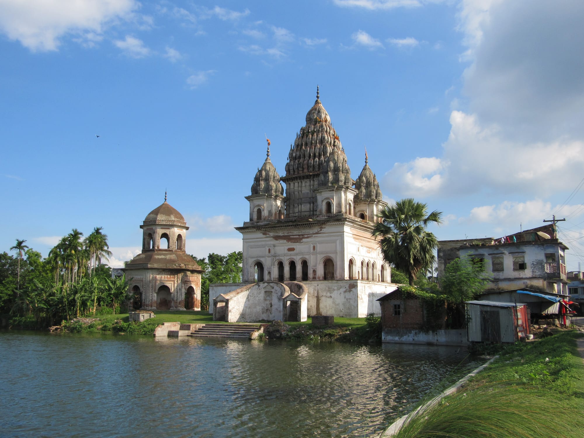 A white Hindu temple with several spired towers beside a pond, with a smaller domed temple and palm trees to its left