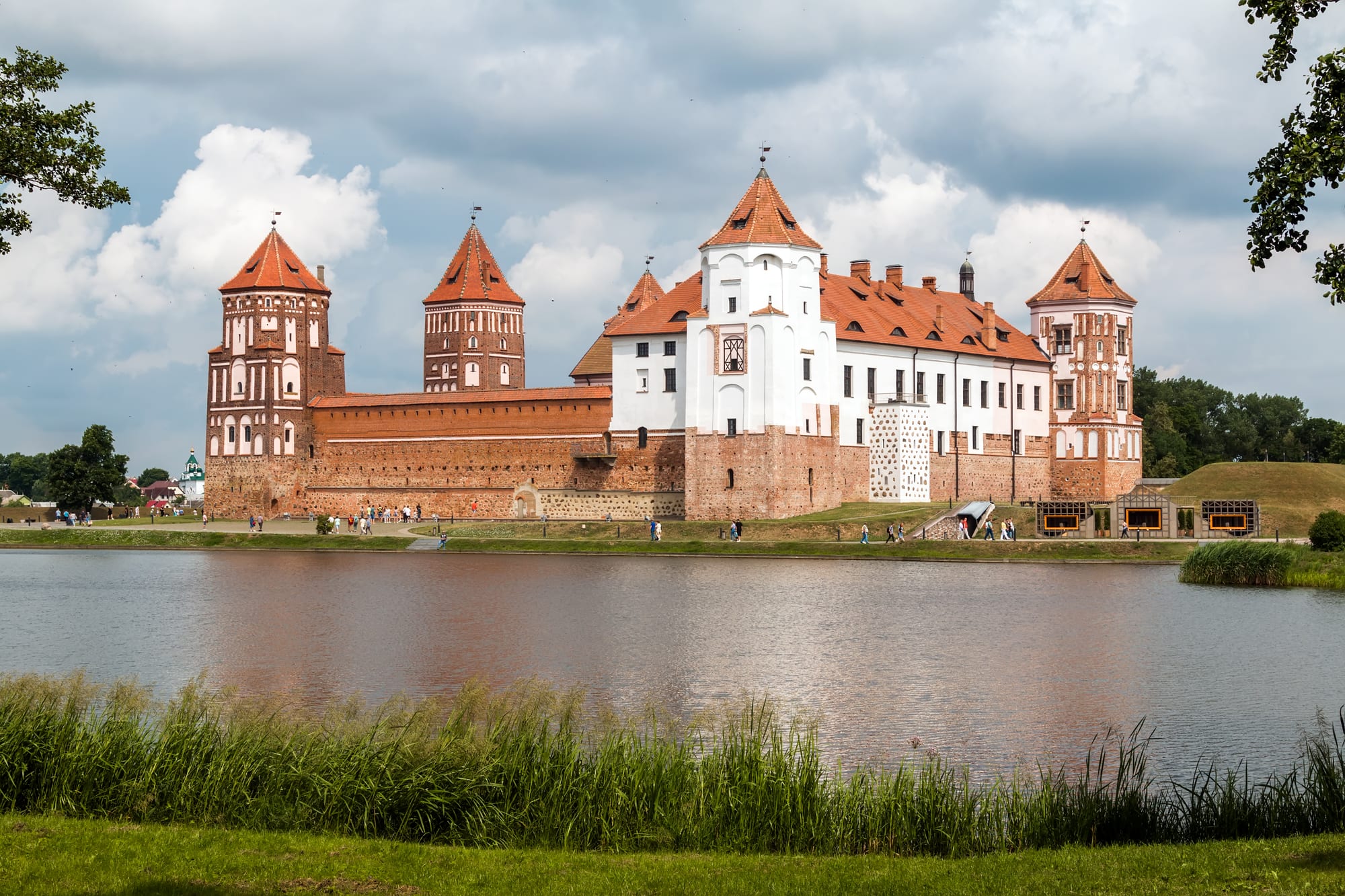 Mir Castle's red-brick towers and white-walled palace wing reflected in a pond under a cloudy summer sky