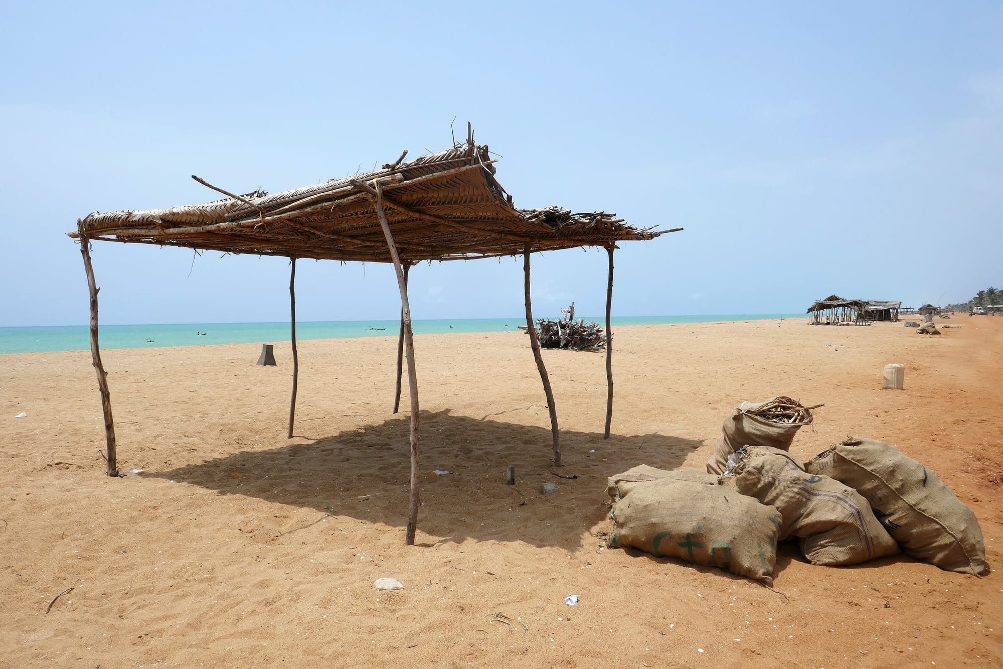 A palm-thatched shelter and sacks on a wide empty beach of orange sand beside a pale turquoise sea
