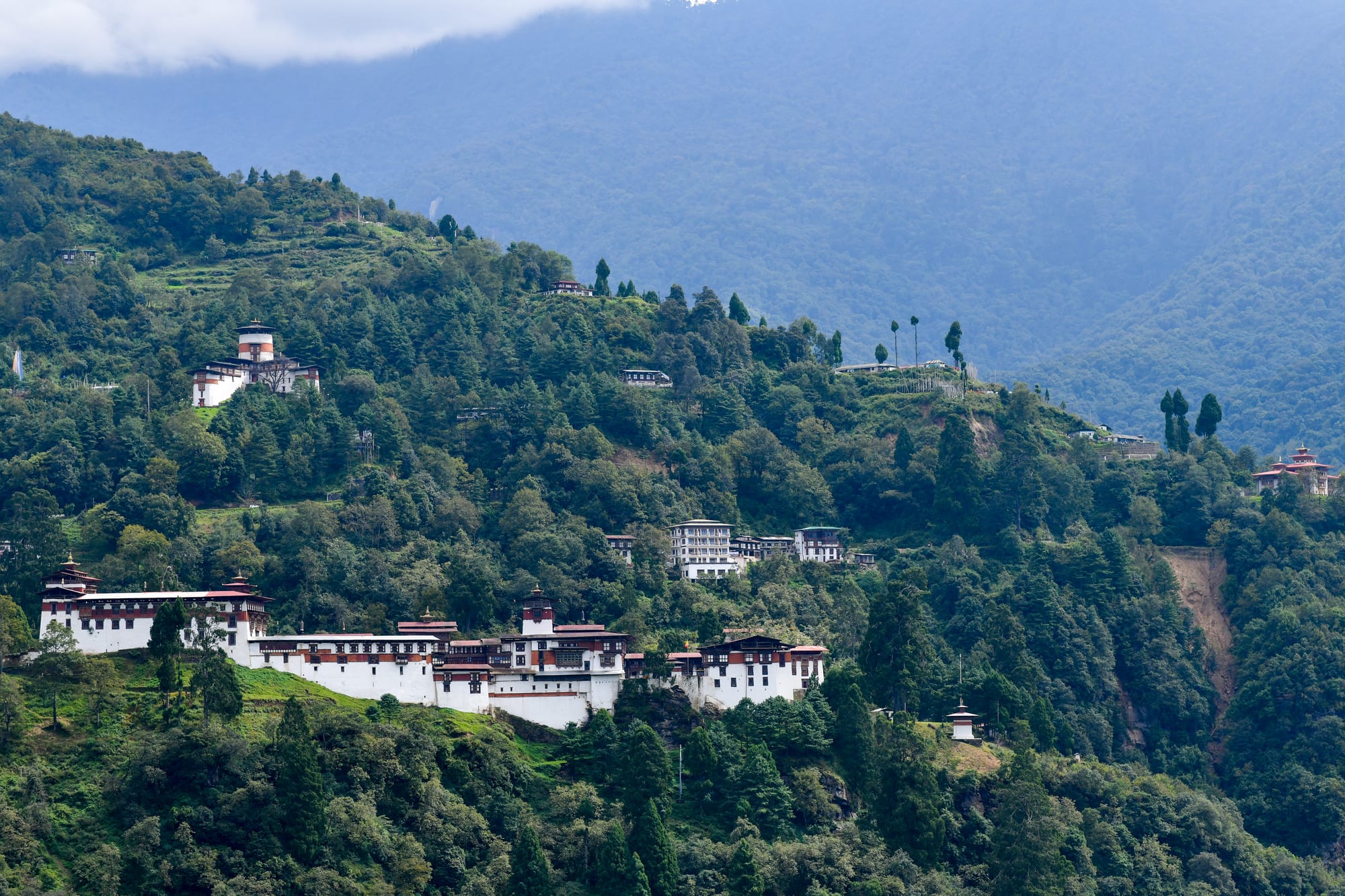 A long white dzong along a forested ridge, with a round watchtower higher on the hillside and cloud over the mountains