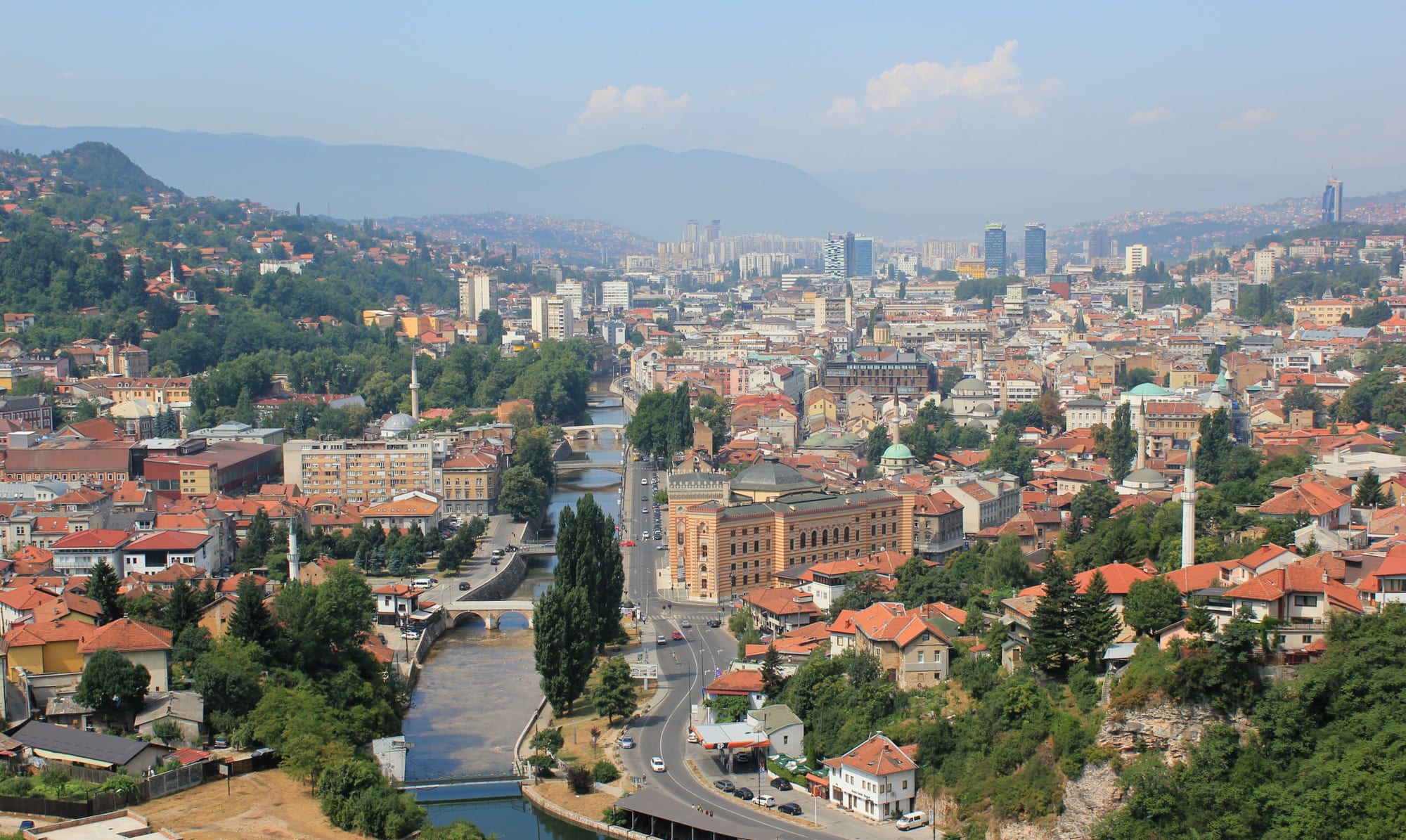 Red roofs, minarets and the striped City Hall of Sarajevo beside the Miljacka river, with hazy mountains beyond