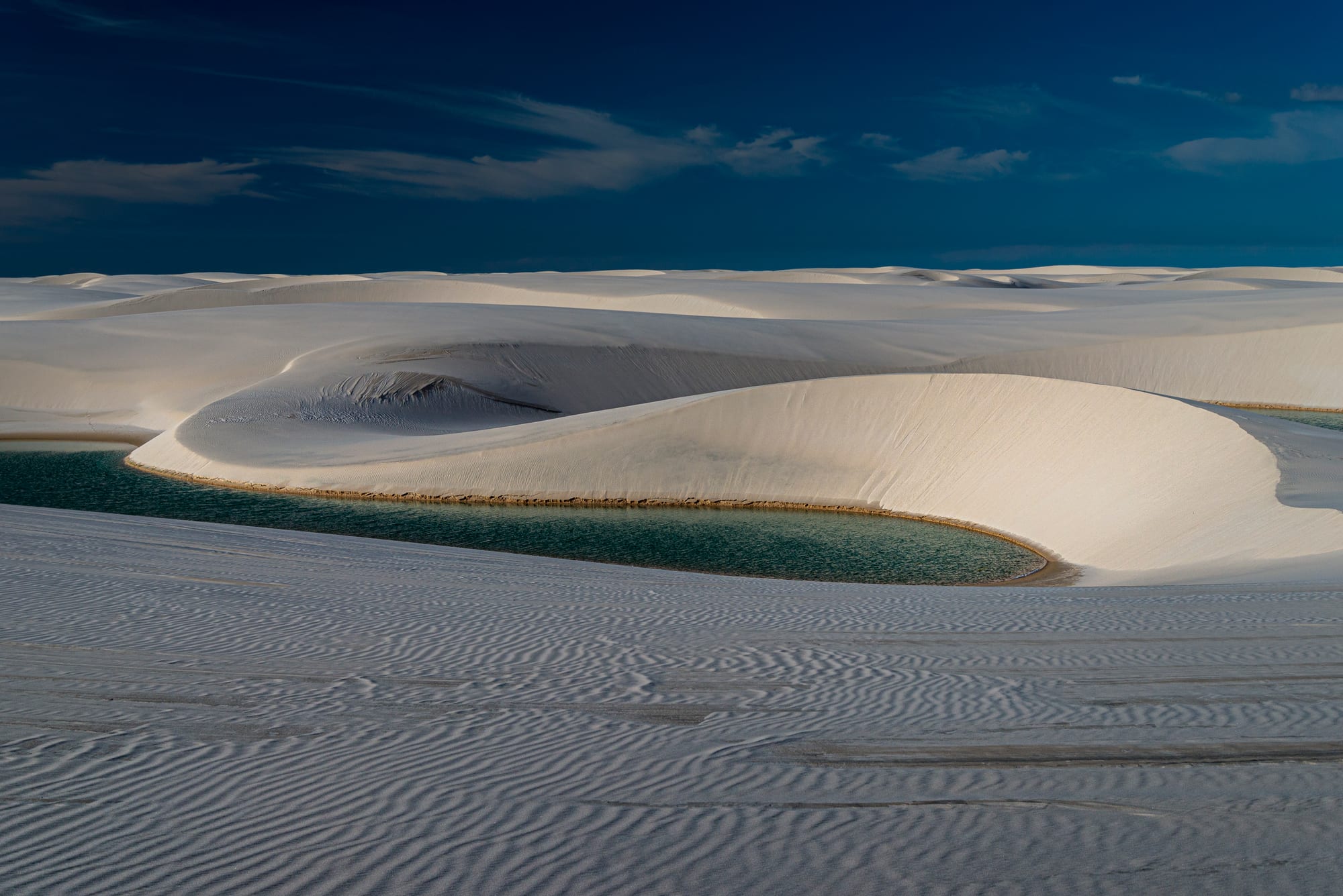 Smooth white sand dunes curving around a green freshwater lagoon under a deep blue sky with thin clouds