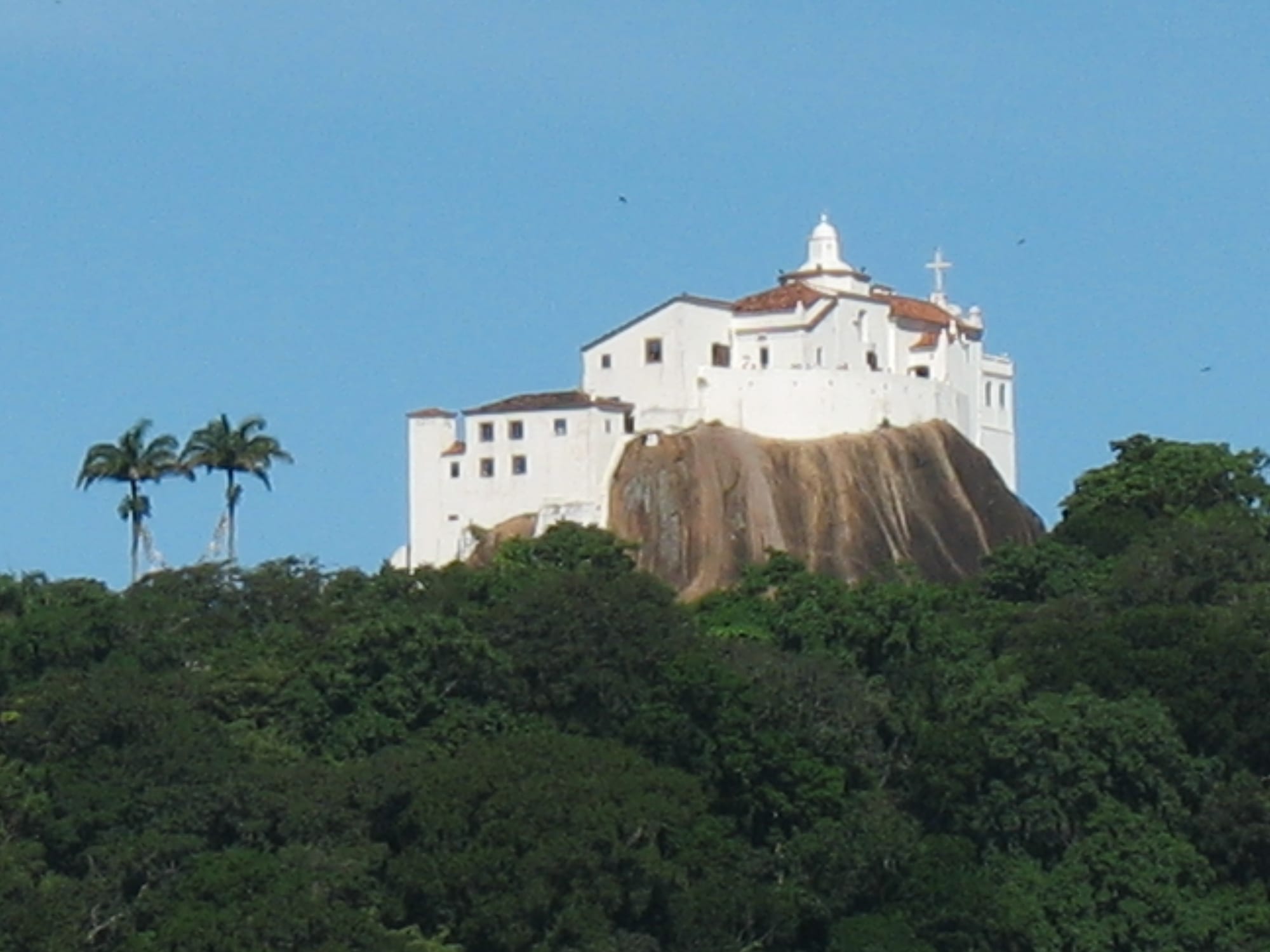 A white colonial convent with a small bell tower and cross perched on a bare granite rock above a forested hill, with two palm trees on the left