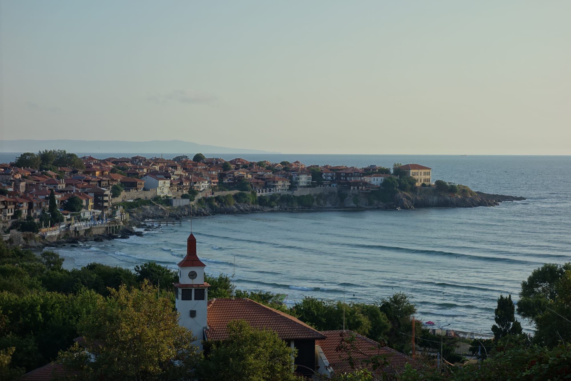 Red-roofed houses of an old town on a rocky peninsula above a calm bay, with a clock tower among trees in the foreground