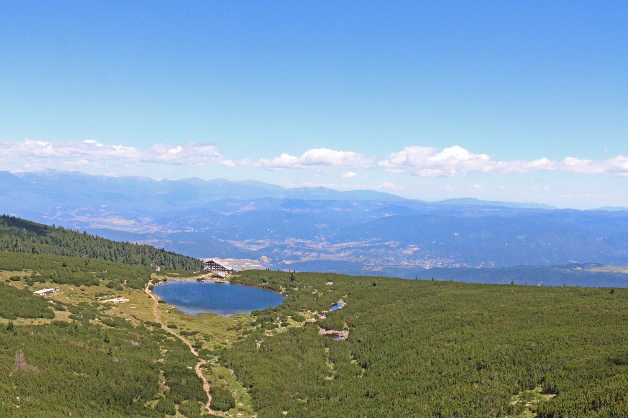 A small blue mountain lake with a hut beside it, set among dwarf pine slopes, with rows of hazy mountain ridges stretching to the horizon