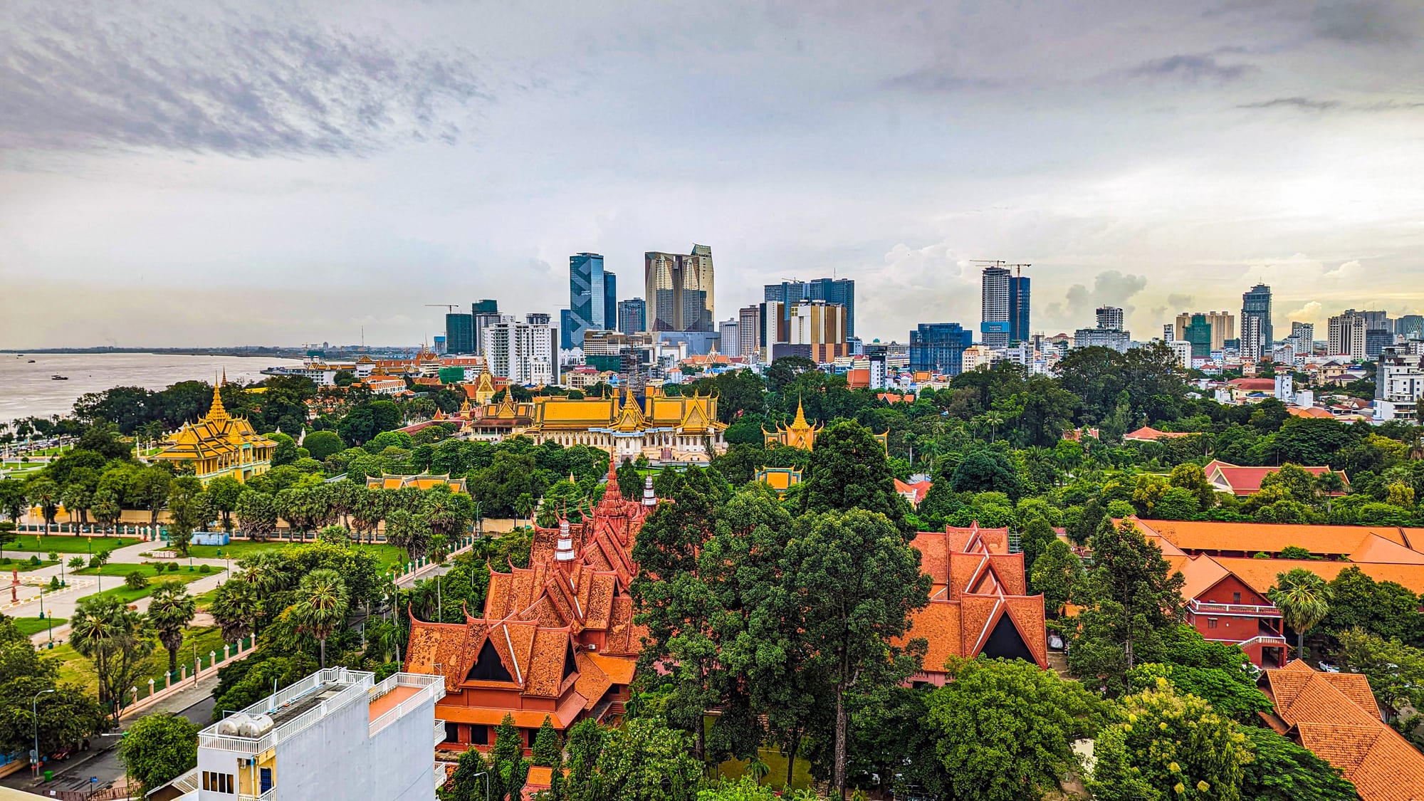Gold-roofed pavilions of the Royal Palace and red-roofed museum among trees, with Phnom Penh's towers and the river behind