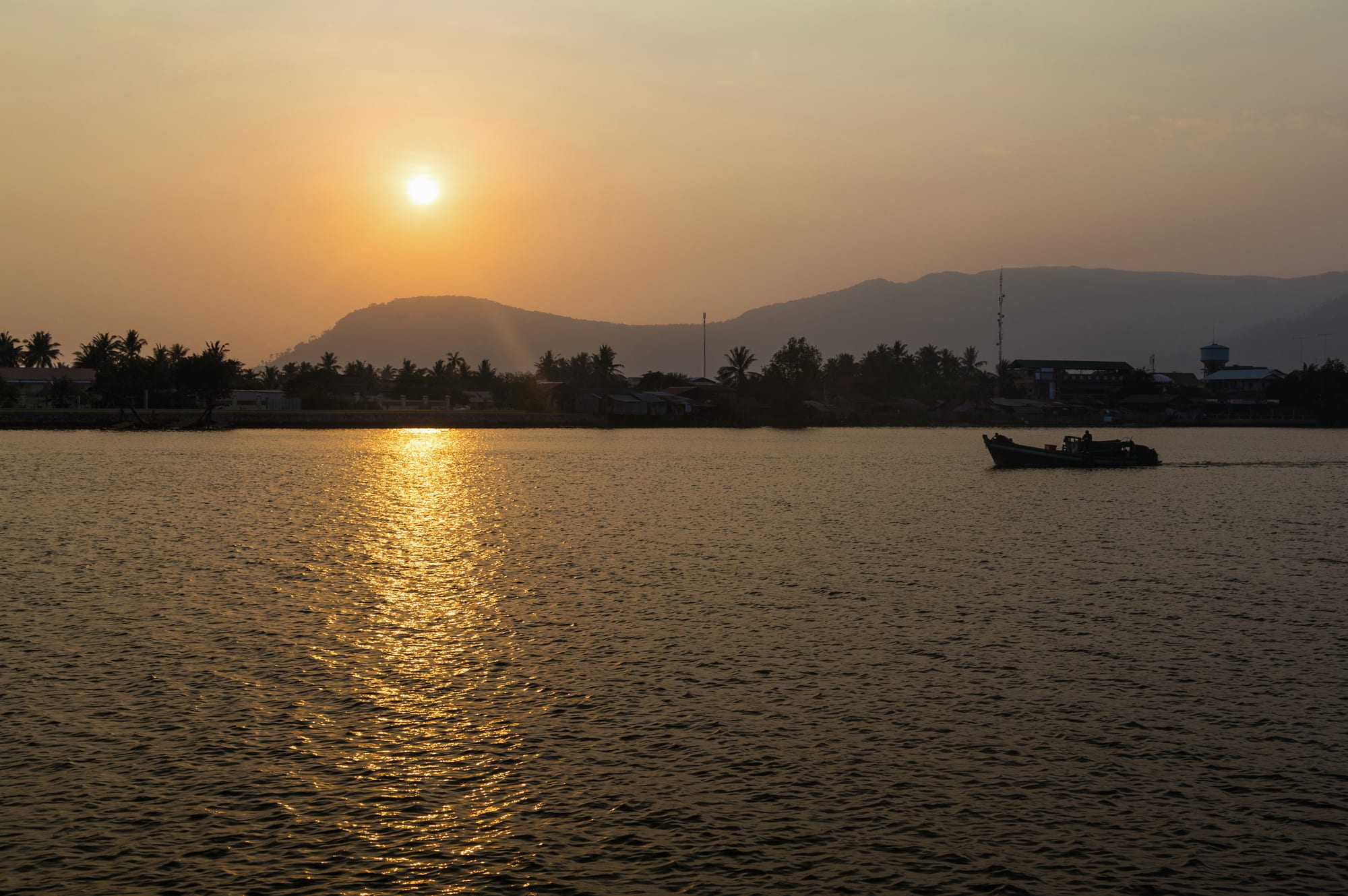 Sunset over a wide calm river with a small boat, palm-lined bank and hazy mountains behind