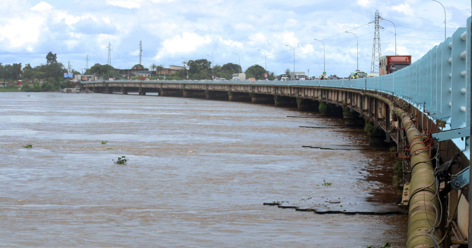 A long concrete road bridge curving across a wide brown river under a cloudy sky, with pylons and trees on the far bank
