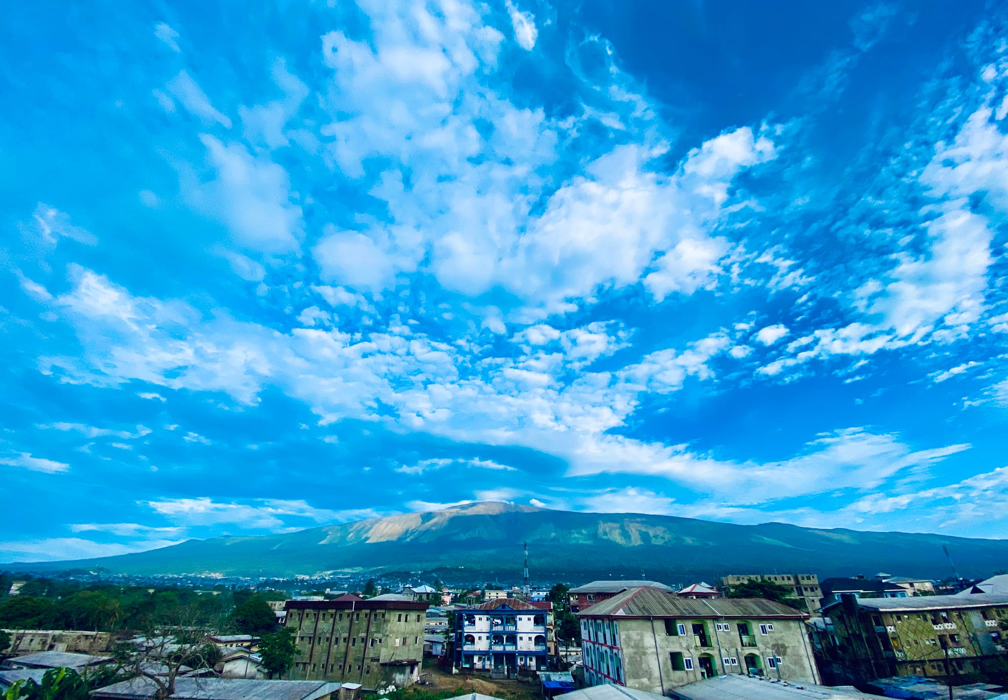 A broad green volcanic mountain under a bright blue sky full of white clouds, with rooftops and apartment blocks of a town in the foreground