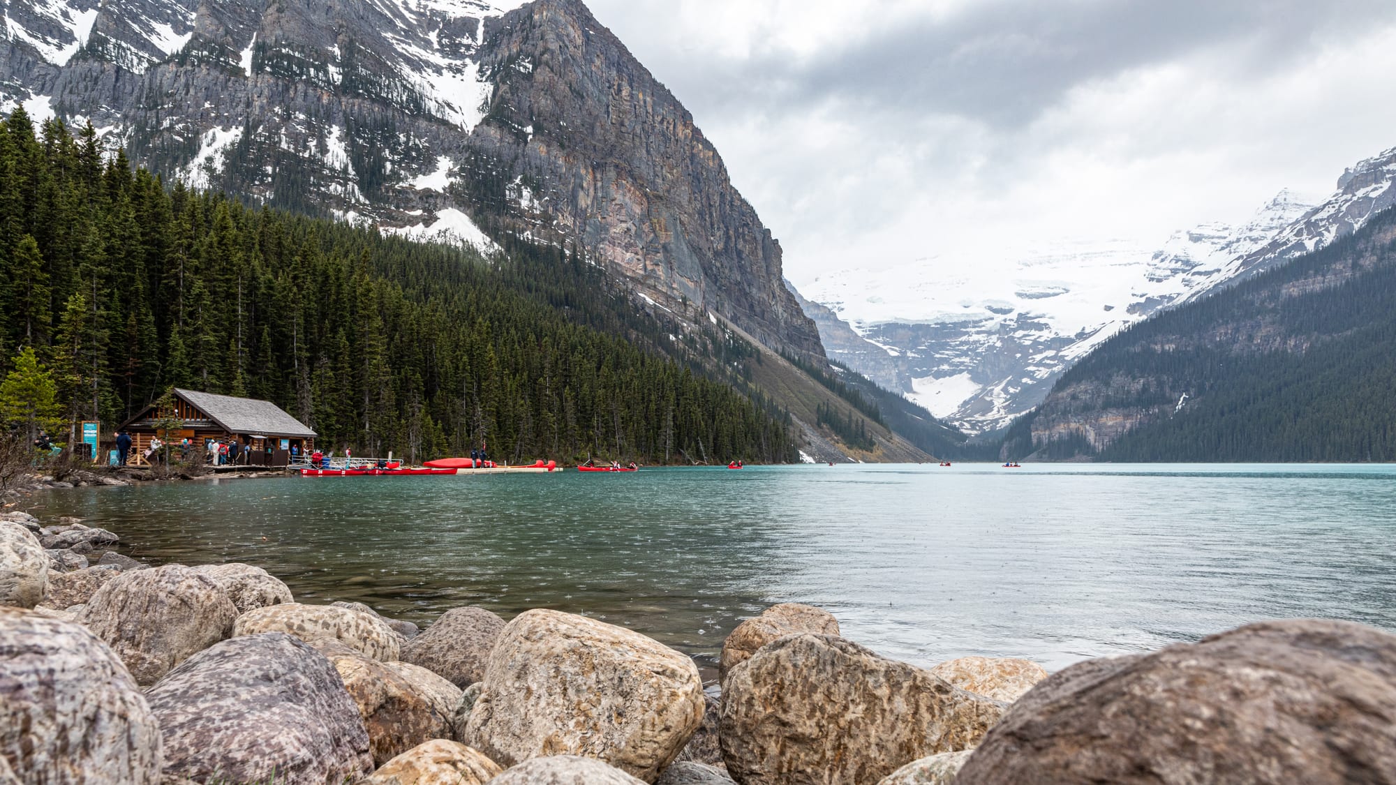 Green-blue Lake Louise under cloud, with a boathouse and red canoes by the forest and a glacier-covered peak at the far end