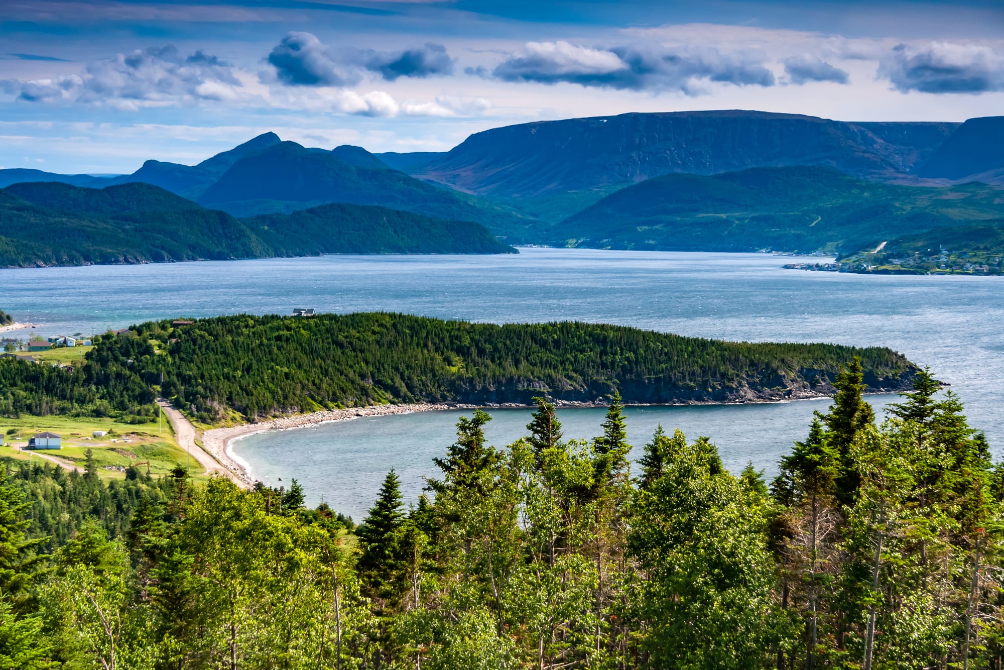 A forested headland and curving beach on a wide blue bay, with flat-topped mountains beyond under broken cloud