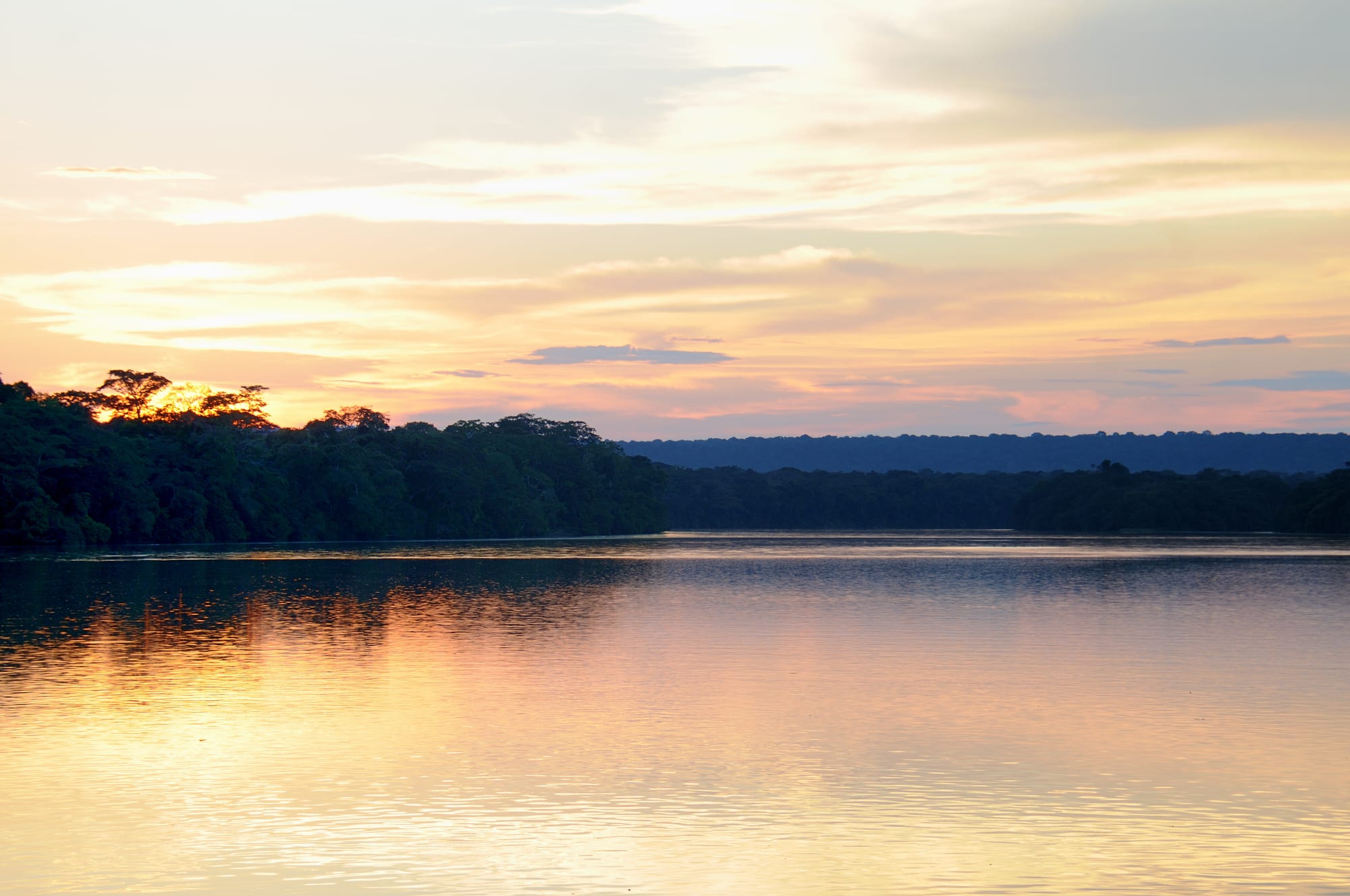 A wide, calm river reflecting a golden sunset sky, with dark rainforest lining both banks