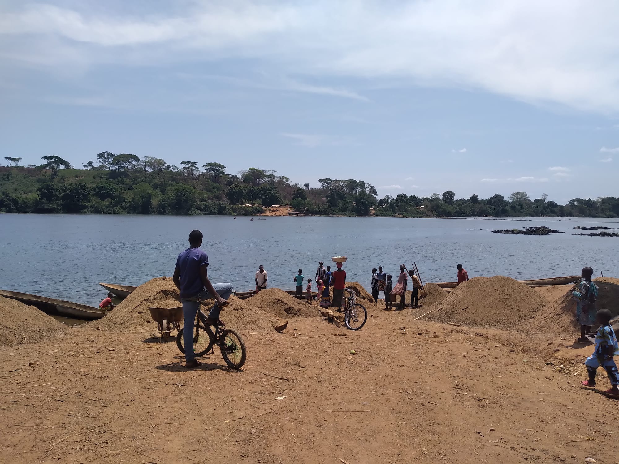 A wide, calm river with a wooded far bank, canoes and piles of sand on the red-earth shore in front