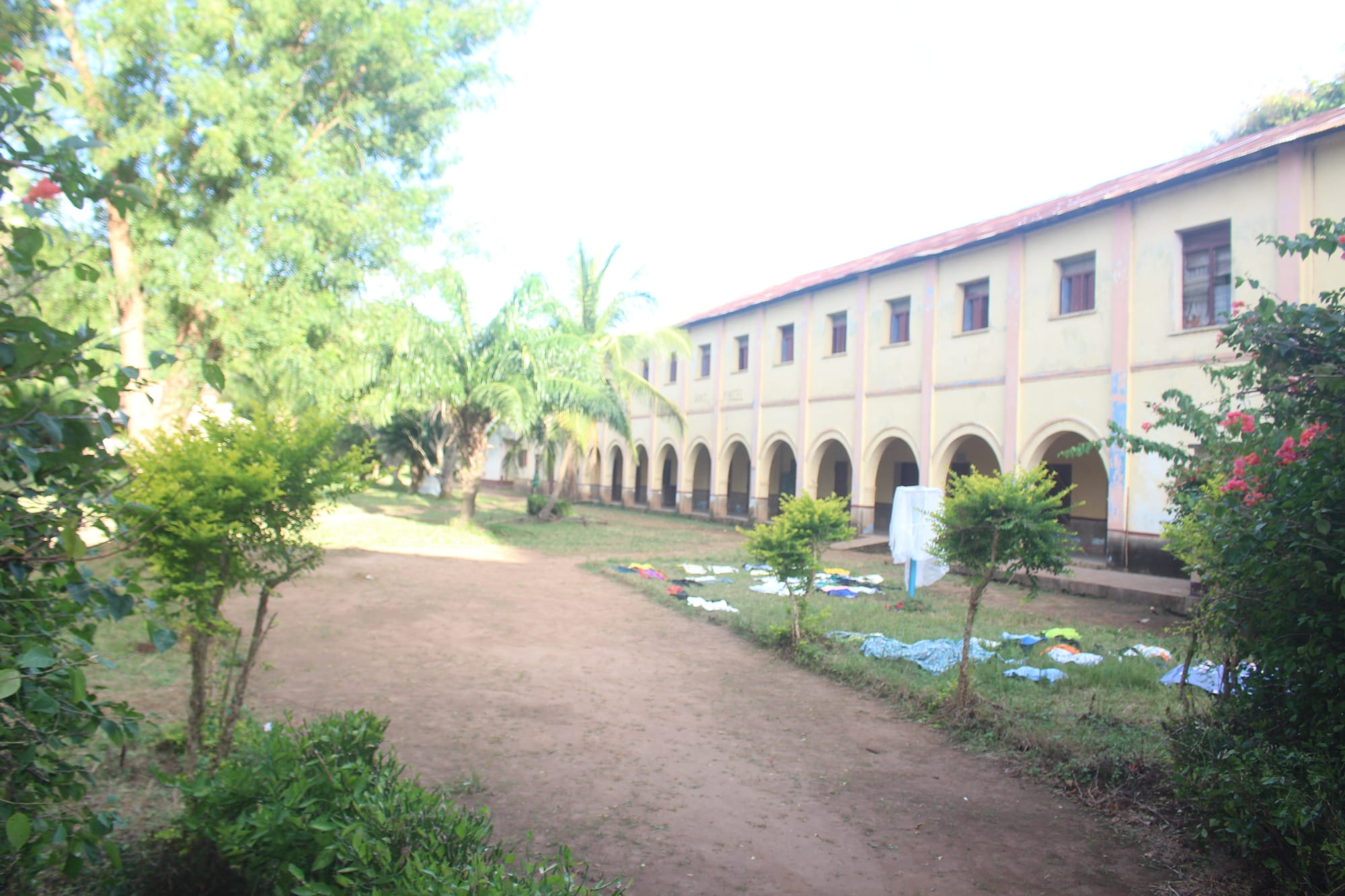A long two-storey arcaded building with a red roof beside a dirt path, trees, palms and laundry drying on the grass