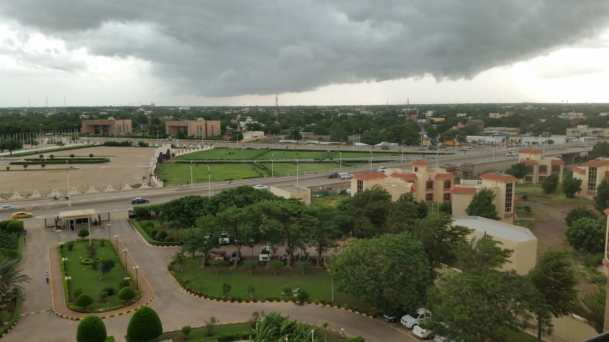 Dark storm clouds over the low green city of N'Djamena, with a wide avenue, lawns and tree-lined streets below