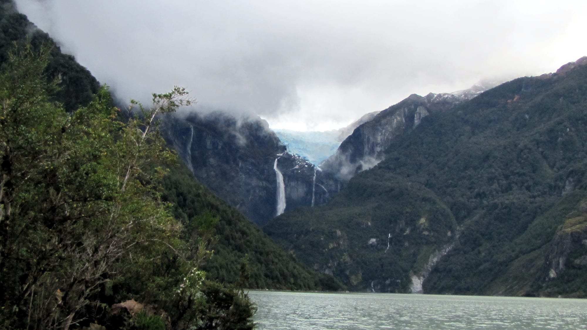 A blue hanging glacier and a long waterfall between steep forested mountains in cloud, above a grey-green lagoon