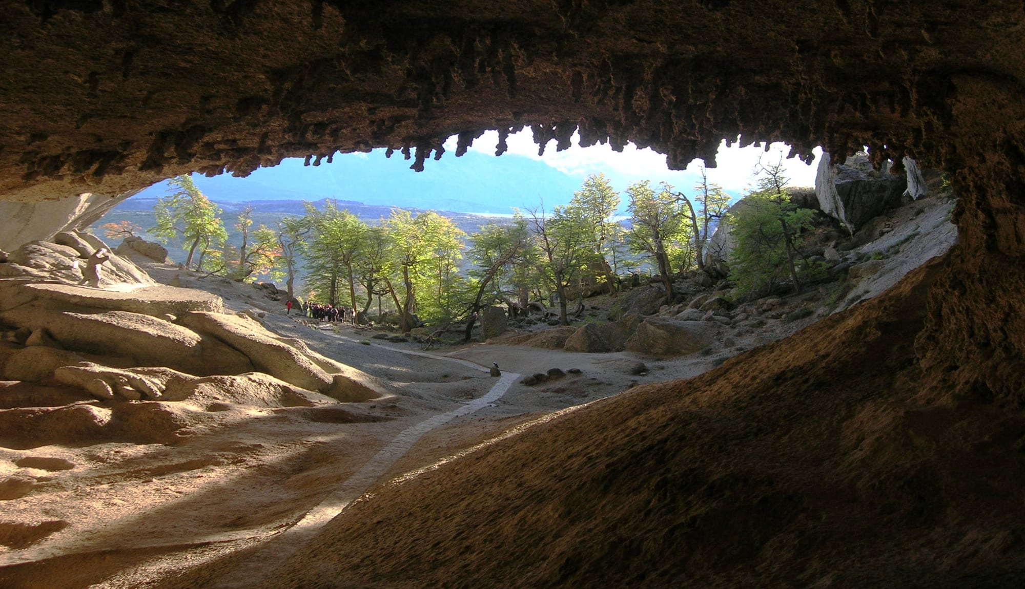 View out from inside a huge cave with a jagged overhanging roof, looking over pale rock and a path toward trees and distant hills