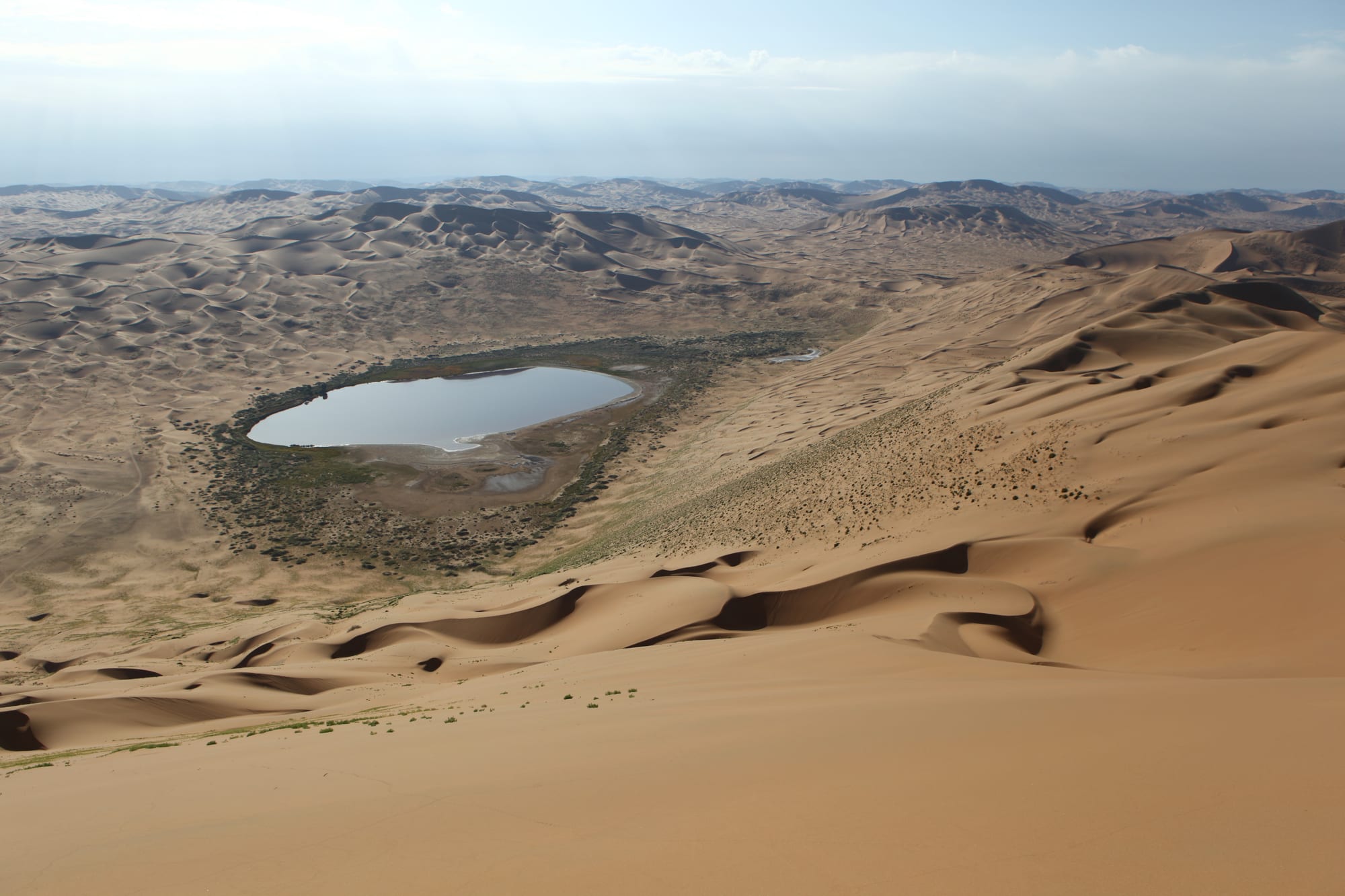 Rippled tan sand dunes stretching to the horizon around a small blue lake fringed with green vegetation