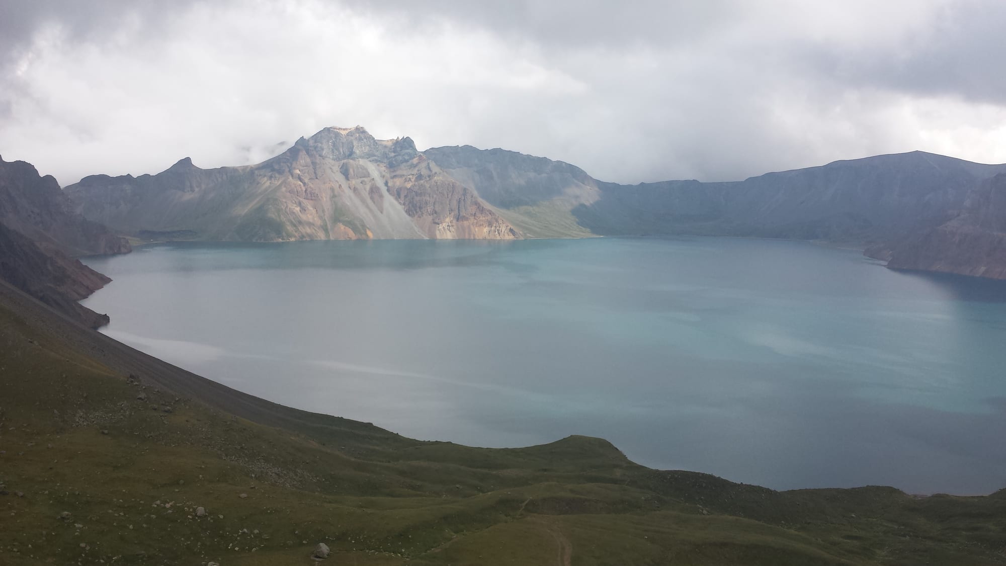 A wide blue-green crater lake ringed by bare grey and ochre volcanic peaks under low cloud, with green slopes in front