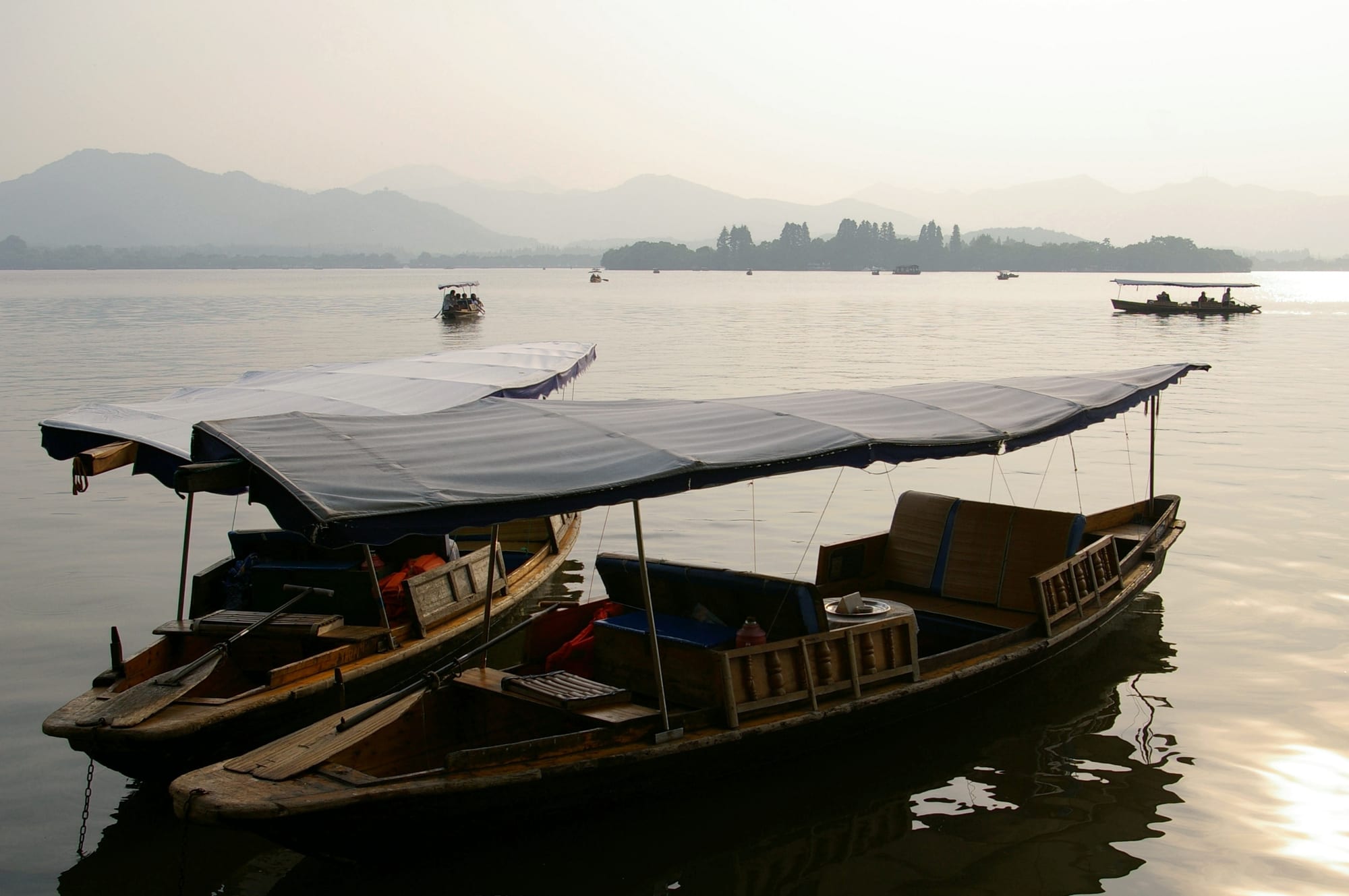 Two canopied wooden boats moored on calm water, with other boats, a wooded island and misty hills beyond