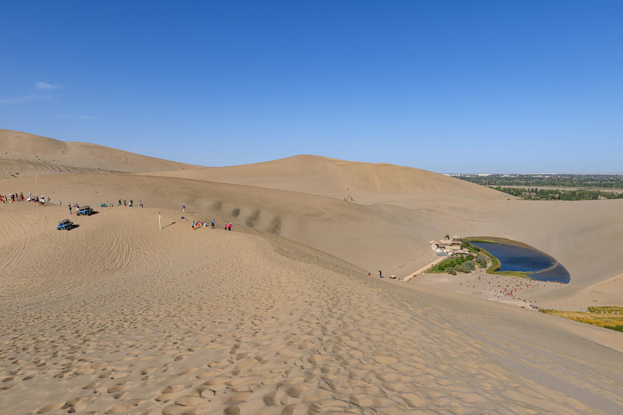 Tall sand dunes under a clear blue sky, with a small crescent-shaped lake and pavilions in a hollow below
