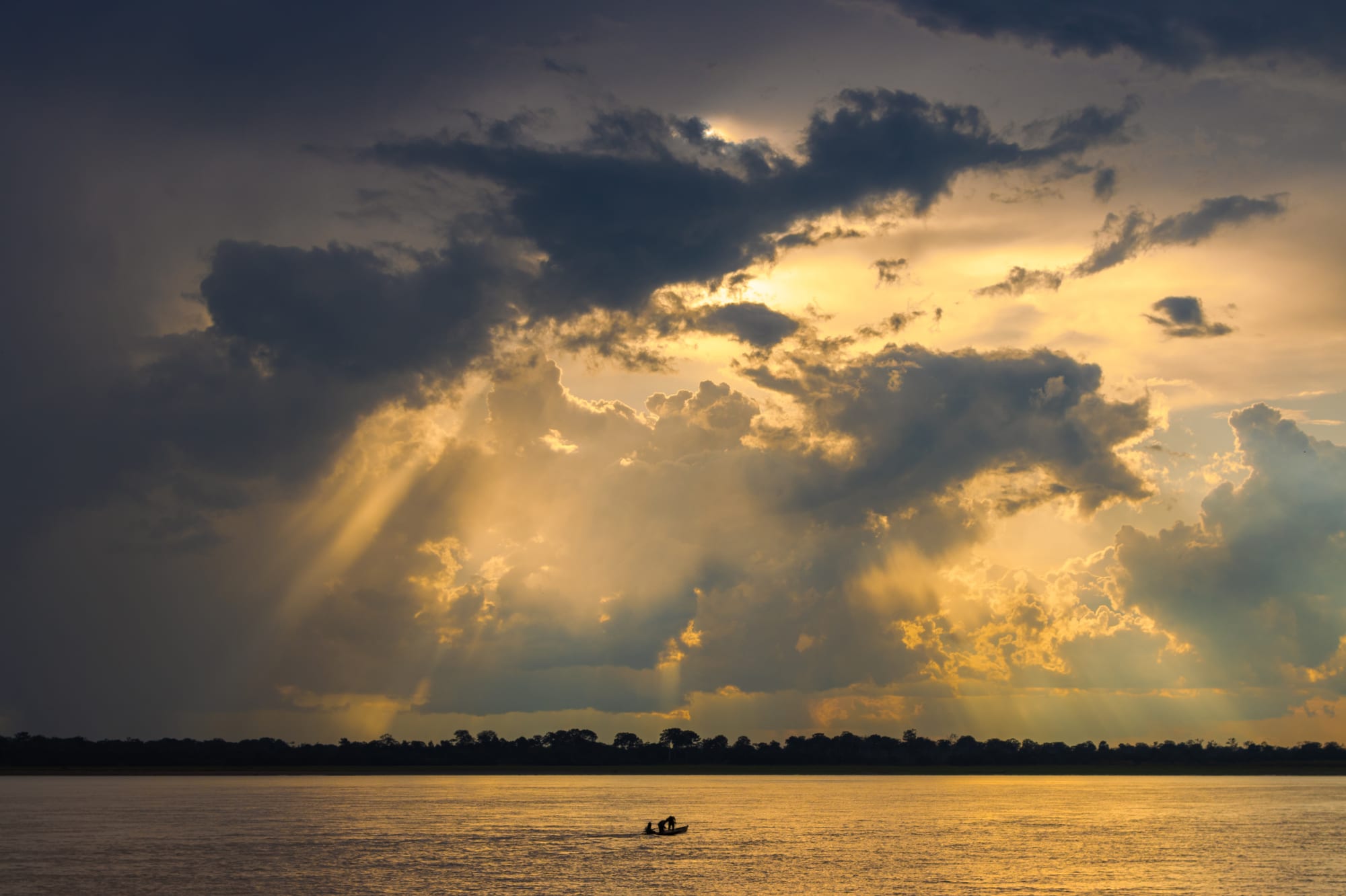 Sun rays breaking through storm clouds over a wide river, with a small fishing boat and a dark line of forest