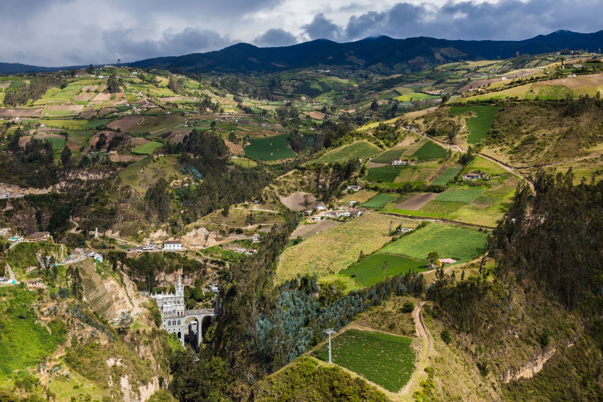 A grey Gothic church on a bridge in a green gorge, below a patchwork of fields and dark mountains