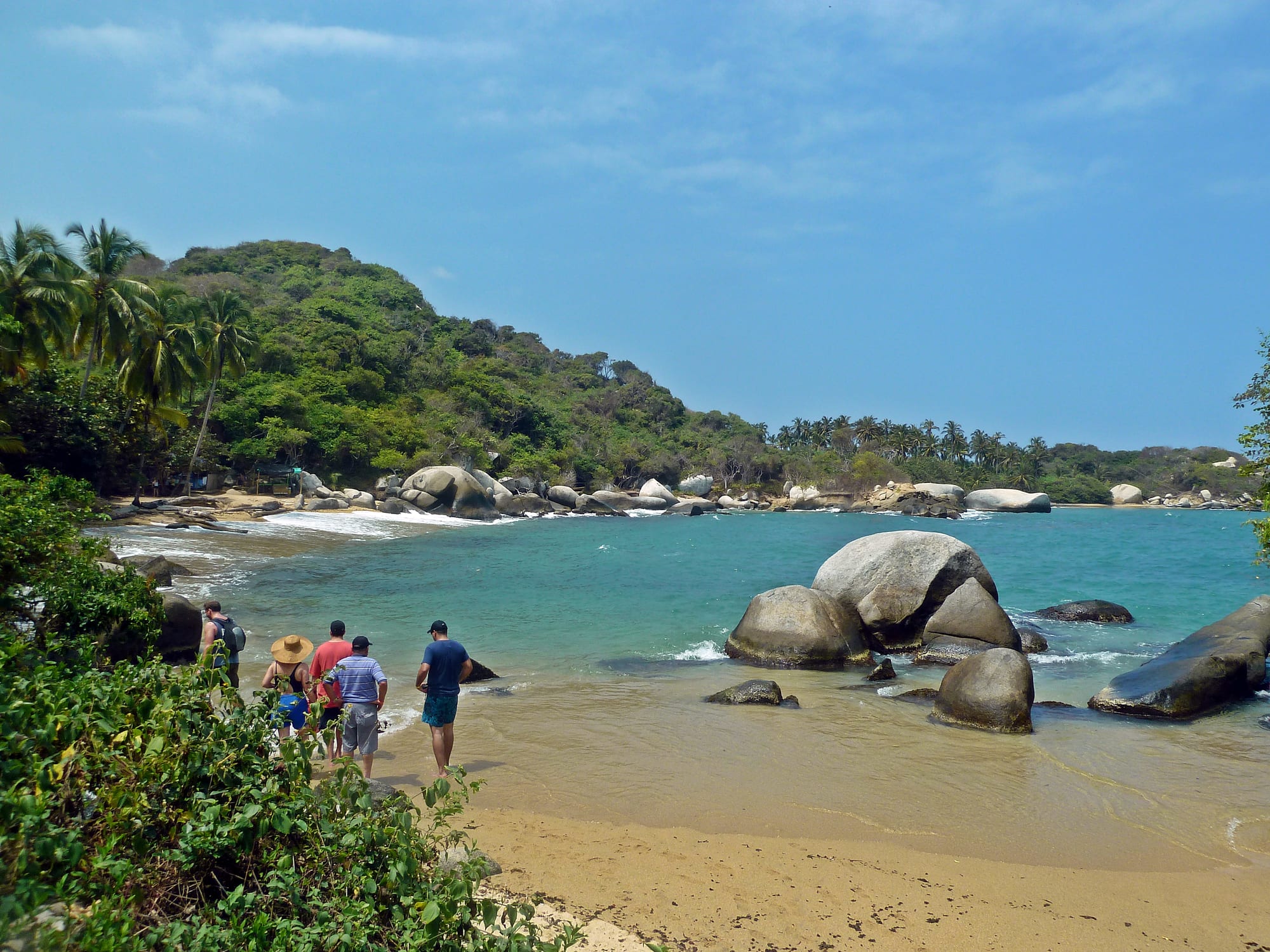 A sandy cove with large granite boulders in turquoise water, backed by palm trees and a forested hill