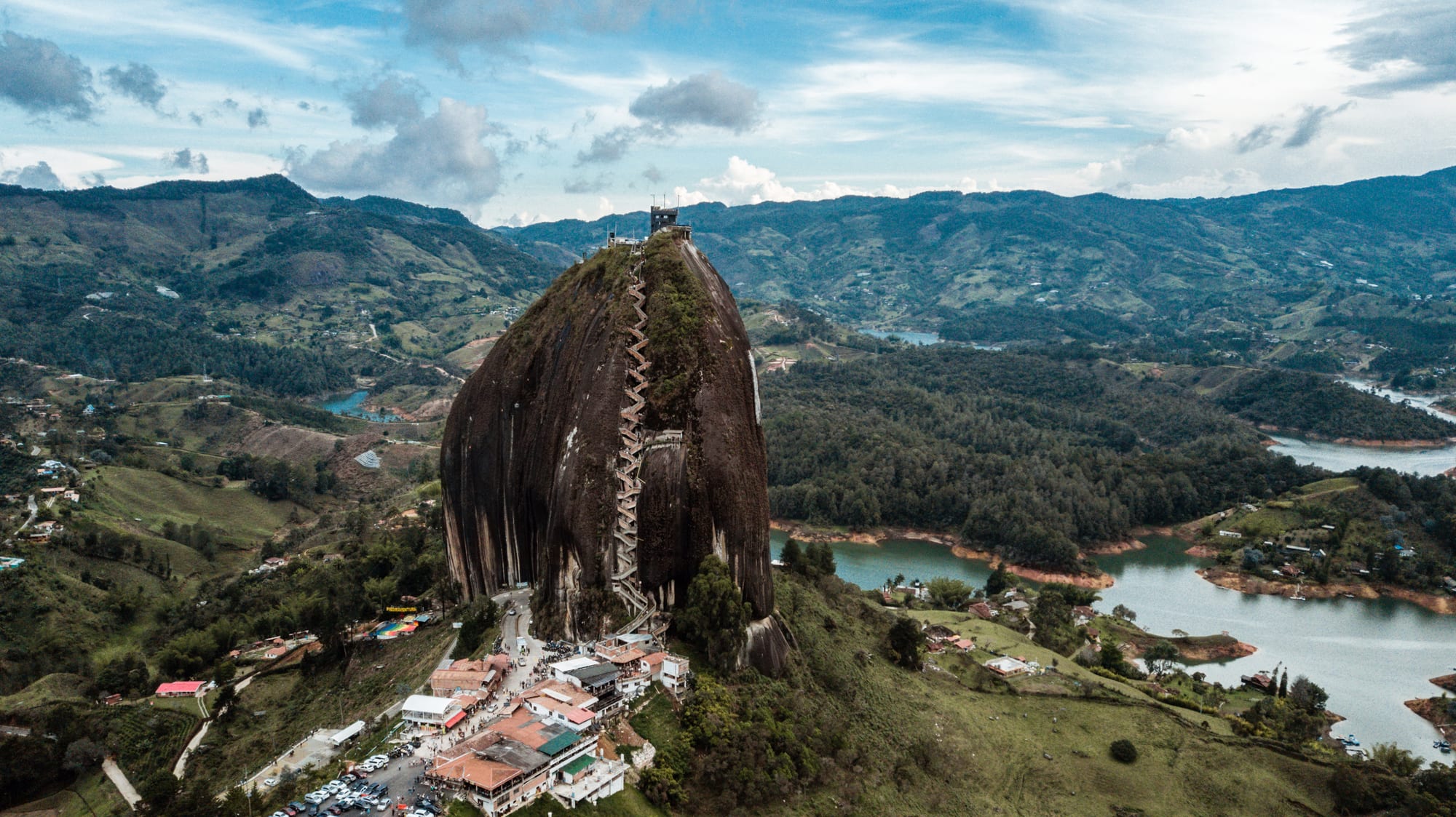 Aerial view of a huge dark granite rock with a zigzag staircase, rising above green hills and winding reservoir arms
