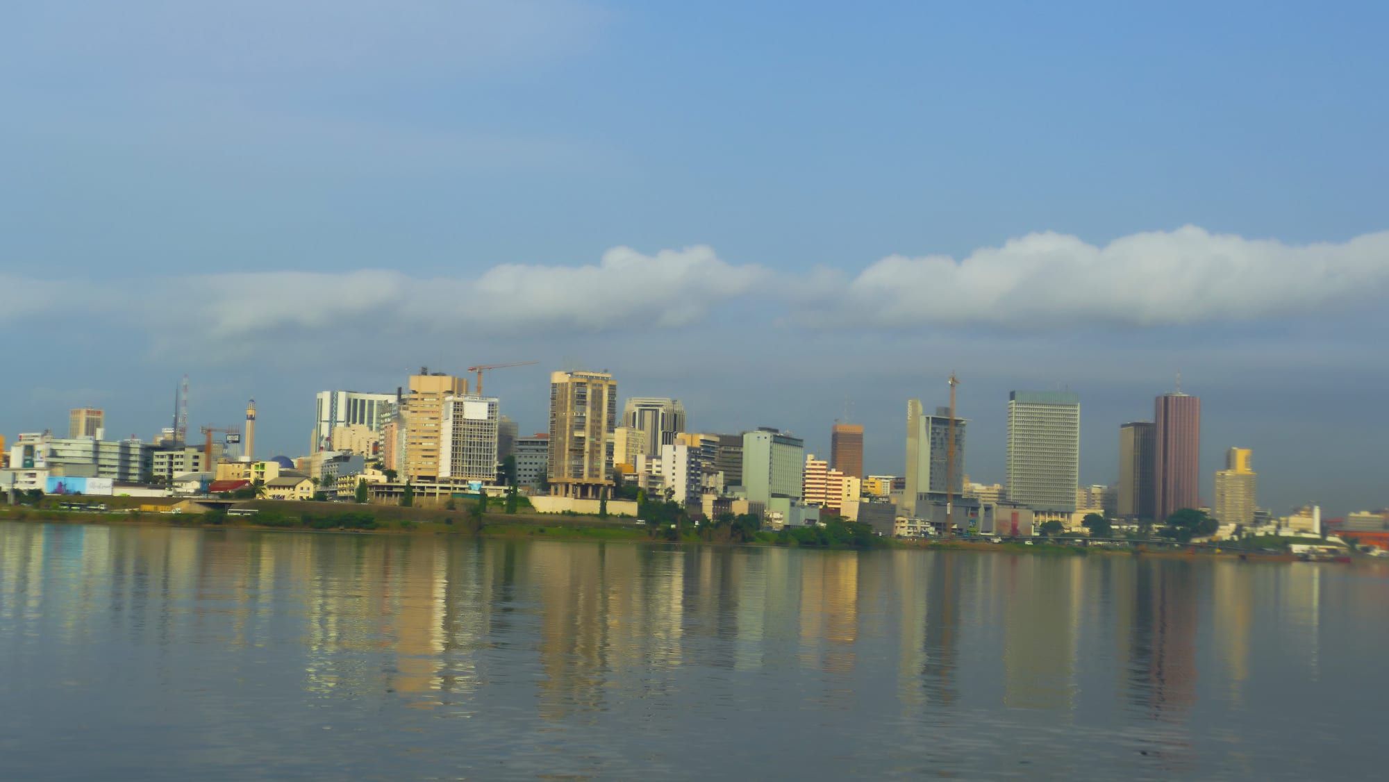 The high-rise towers of Abidjan's Plateau district reflected in the calm water of the Ébrié lagoon under a blue sky